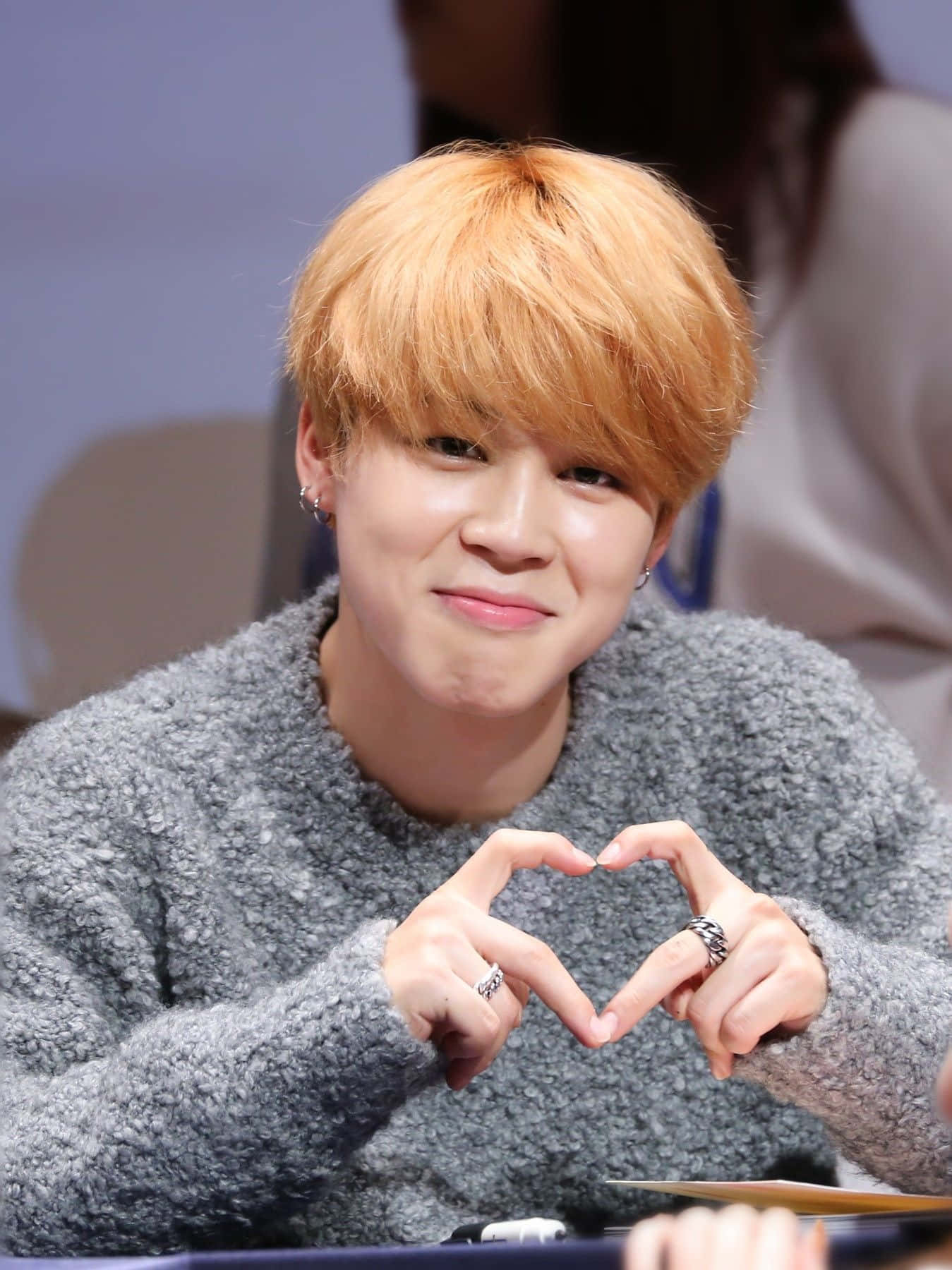 BTS 桌布 壁紙 — 一位橙髮年輕男子戴著戒指，雙手比出心形，溫暖地微笑。