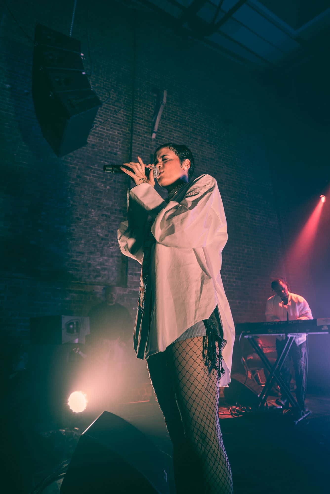 kehlani 桌布 壁紙 — 一位歌手在燈光昏暗、磚牆環繞的舞台上，在戲劇性的聚光燈下投射出彩色光束，熱情地表演。