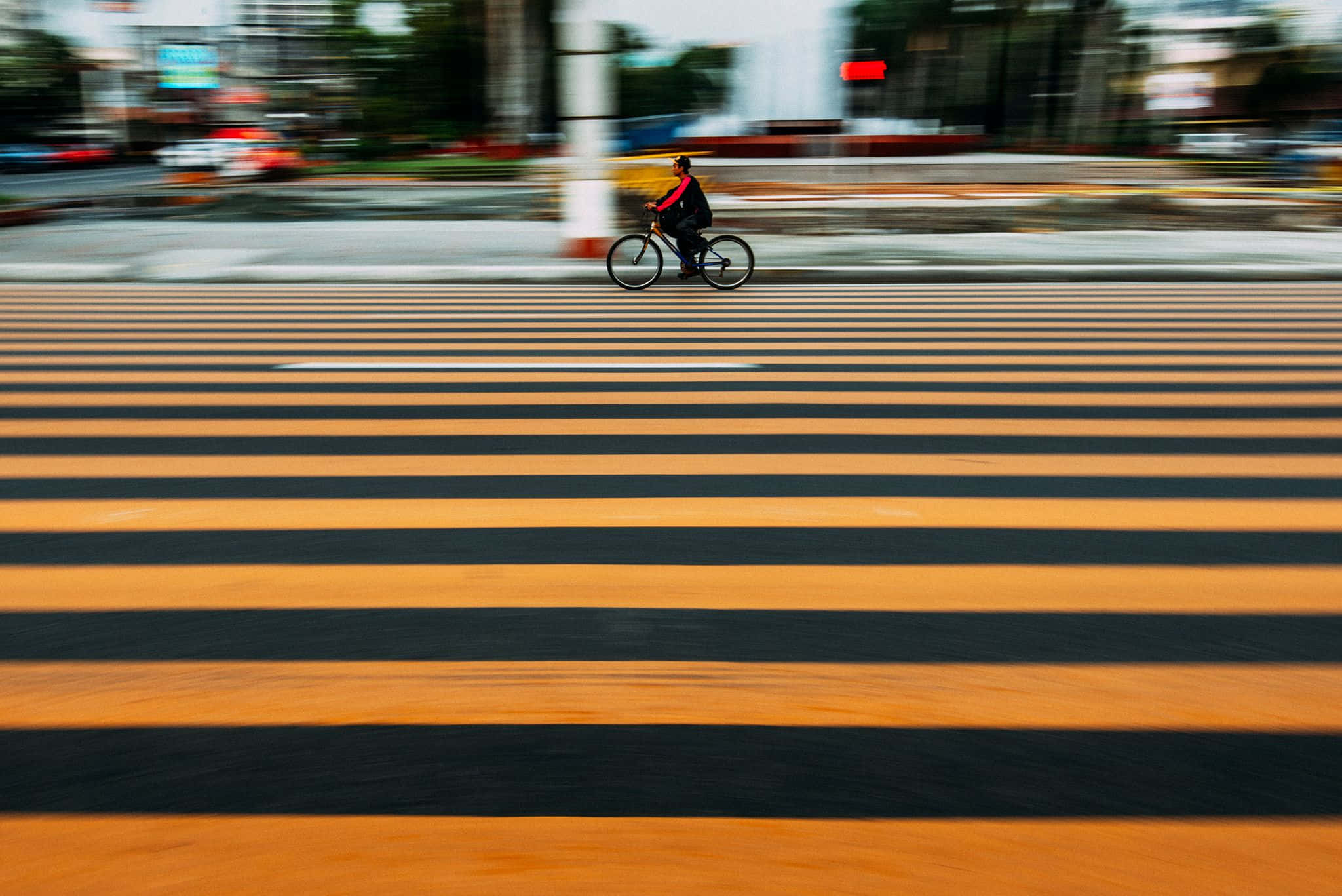橘色 桌布 壁紙 — 一位正在移動的自行車騎士穿過一條大膽的橙色和黑色條紋行人穿越道，背景是模糊的城市景觀。