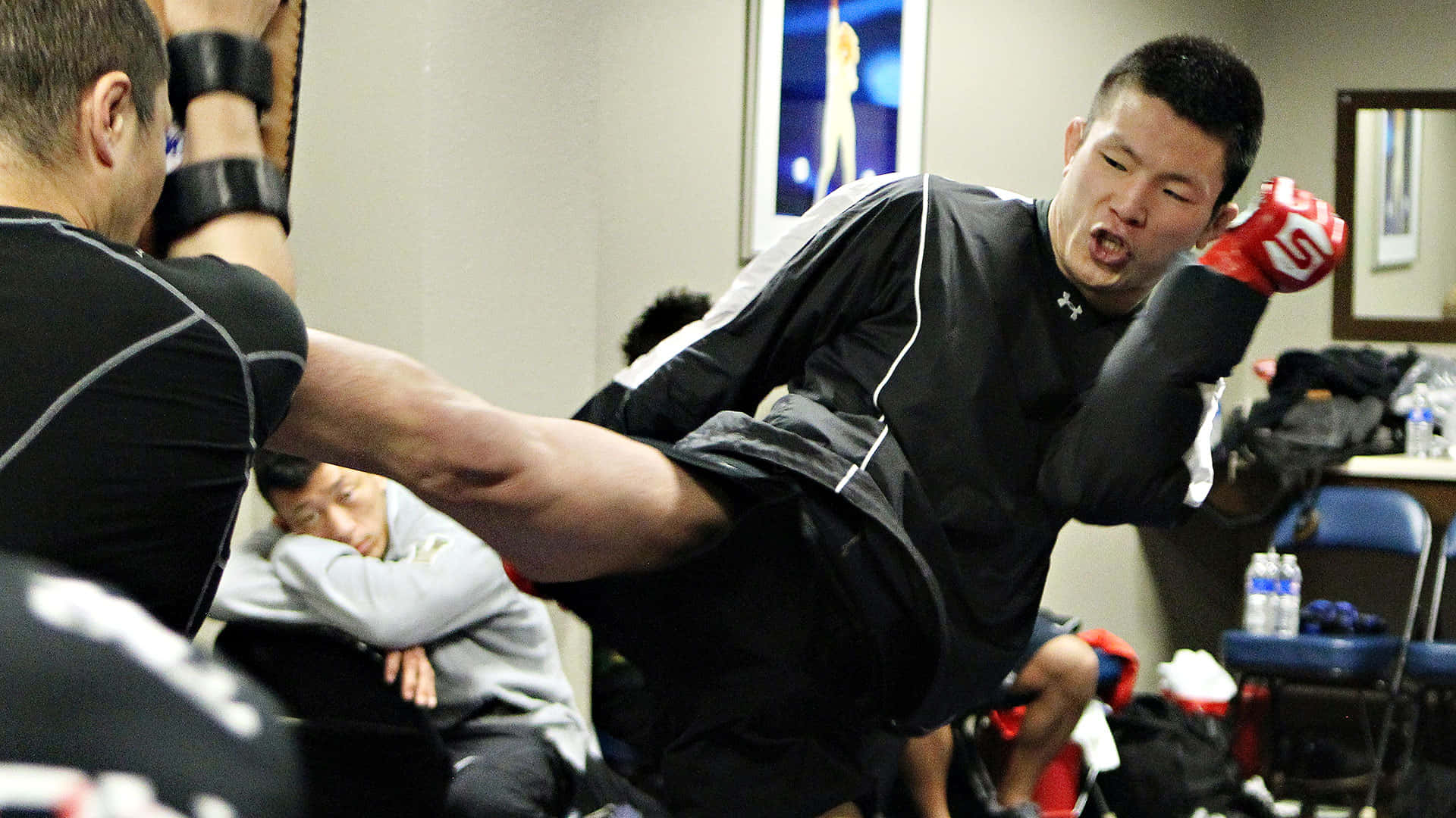 martial arts 桌布 壁紙 — 一位武術家在健身房施展強勁的高踢，教練阻擋，另一名學員觀看。