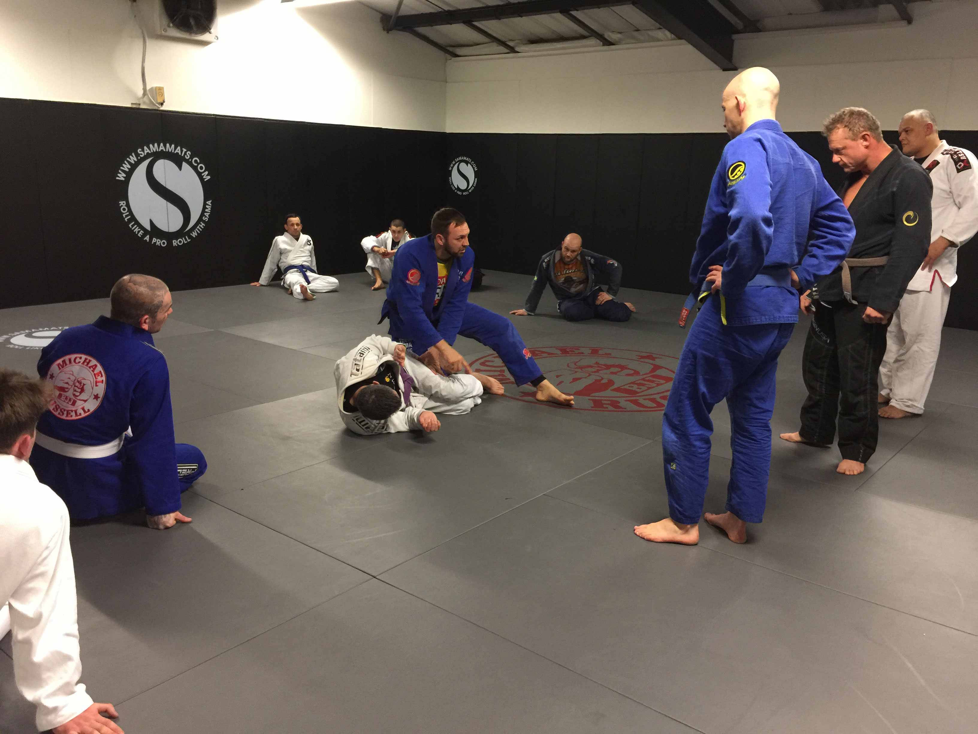 martial arts 桌布 壁紙 — 一位武術教練向學生演示纏鬥技巧，其他學生在黑色牆壁和灰色墊子的健身房中觀看。