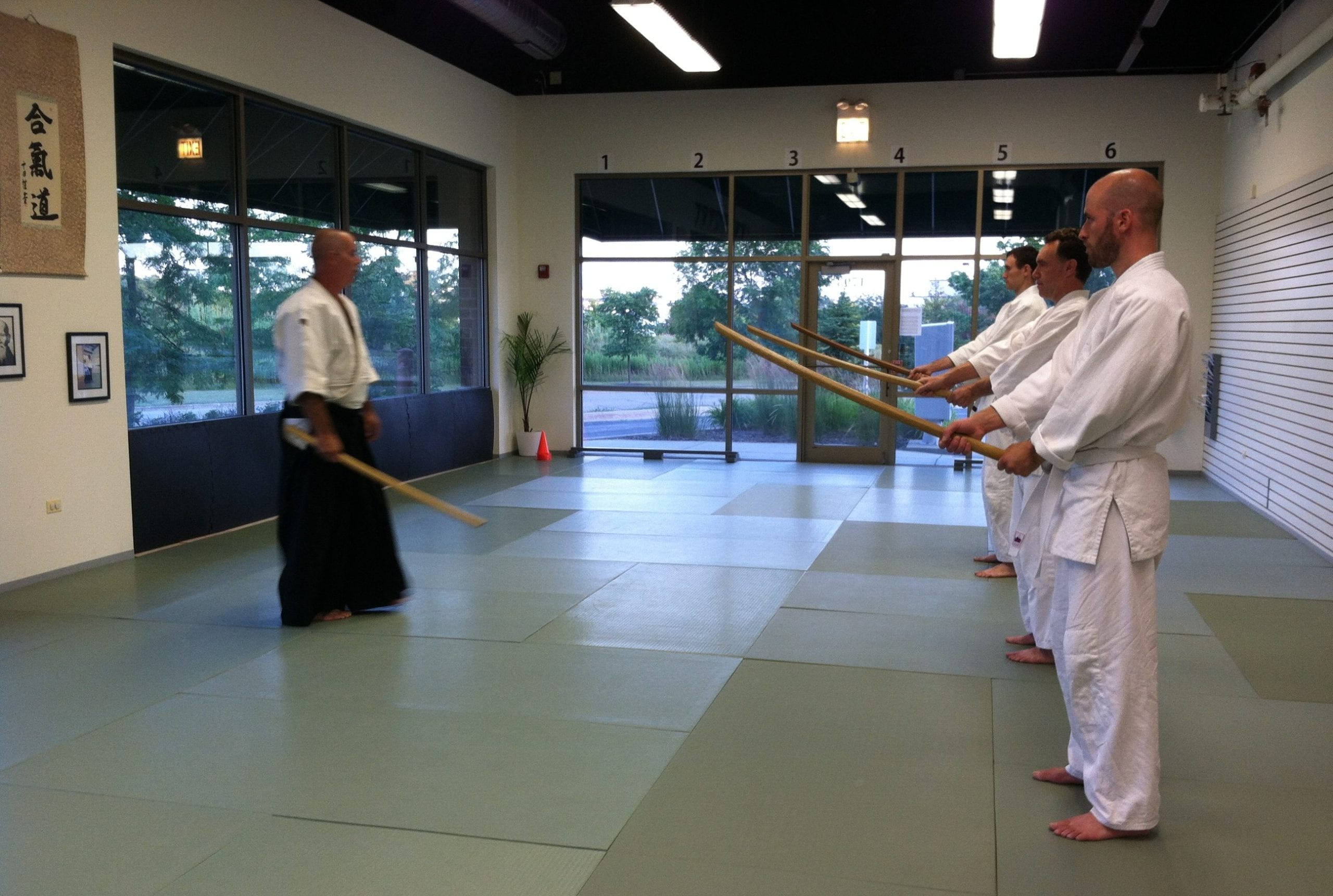 martial arts 桌布 壁紙 — 一位武術教練用木劍示範，身穿白色制服的學生在有大窗戶的道場練習。