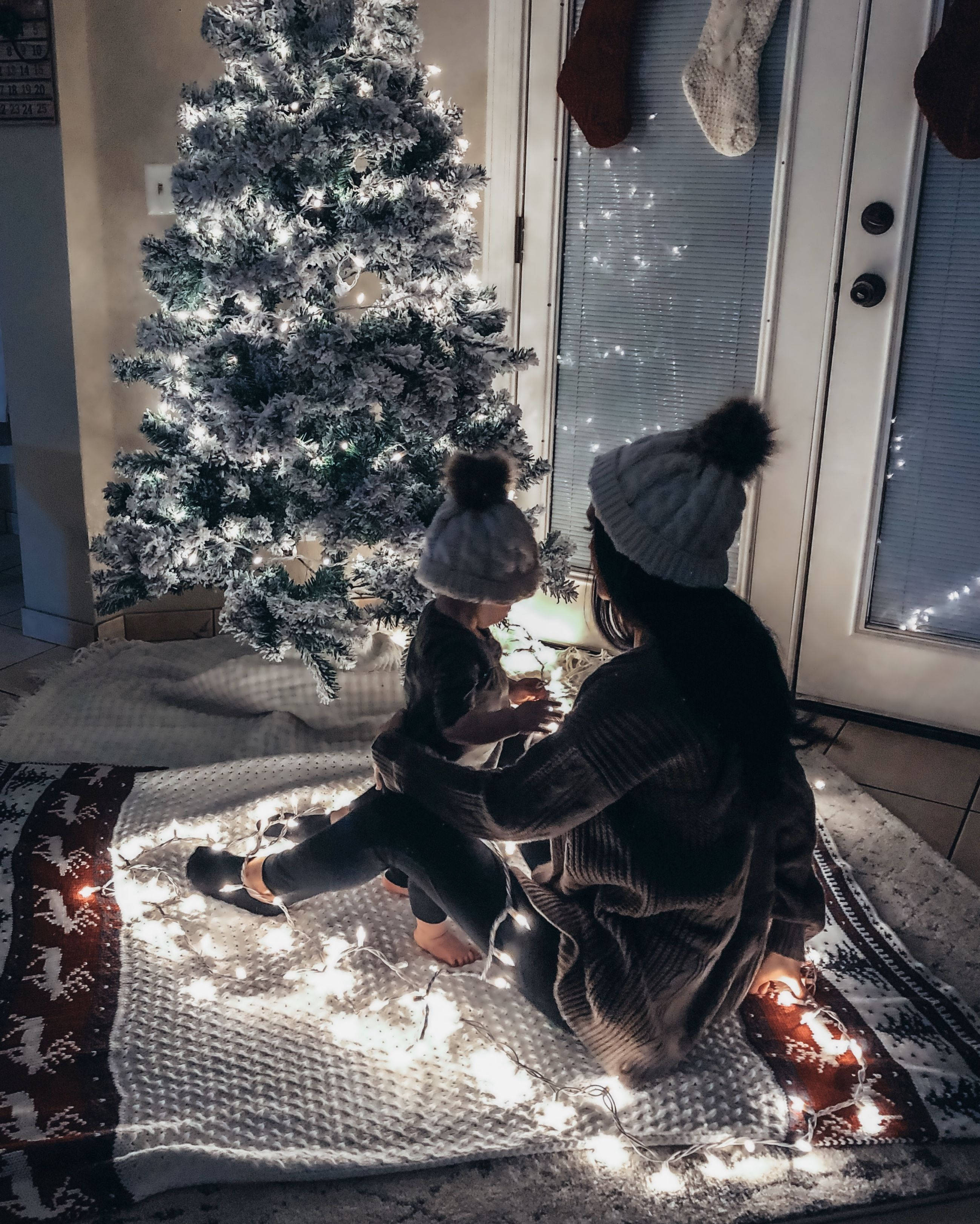 Cozy 桌布 壁紙 — 一位母親和一個孩子一起坐在有圖案的地毯上，在一棵植絨聖誕樹下，周圍環繞著閃爍的童話燈。