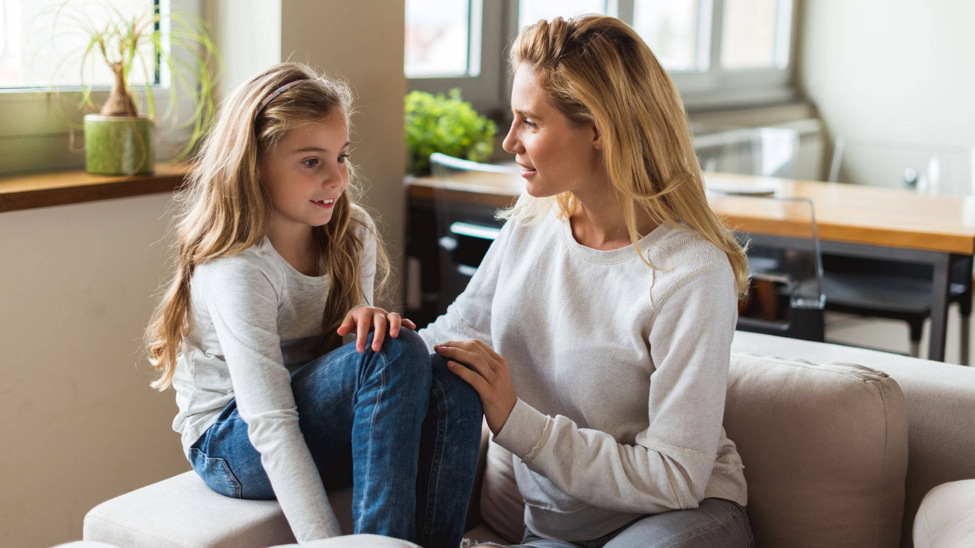 Mom 桌布 壁紙 — 一位母親和女兒在明亮的現代客廳中，一起坐在米色沙發上分享溫馨時刻。