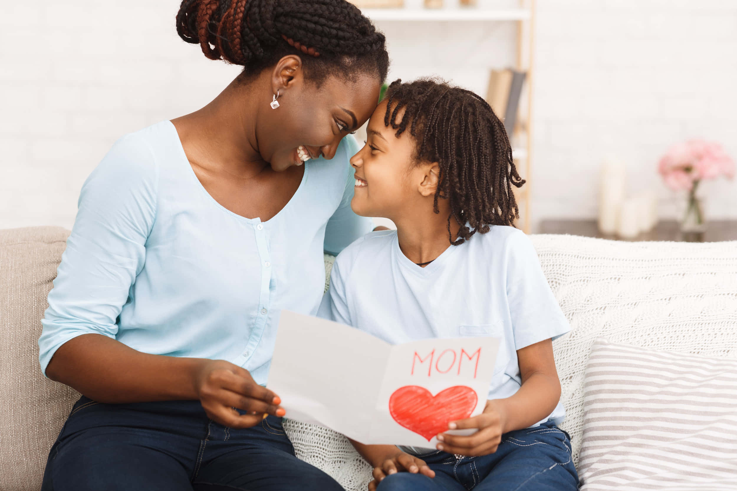 Mother Love 桌布 壁紙 — 一位母親和女兒在沙發上分享溫馨時刻，孩子遞上一張自製卡片，上面有紅色心形和「媽媽」字樣。