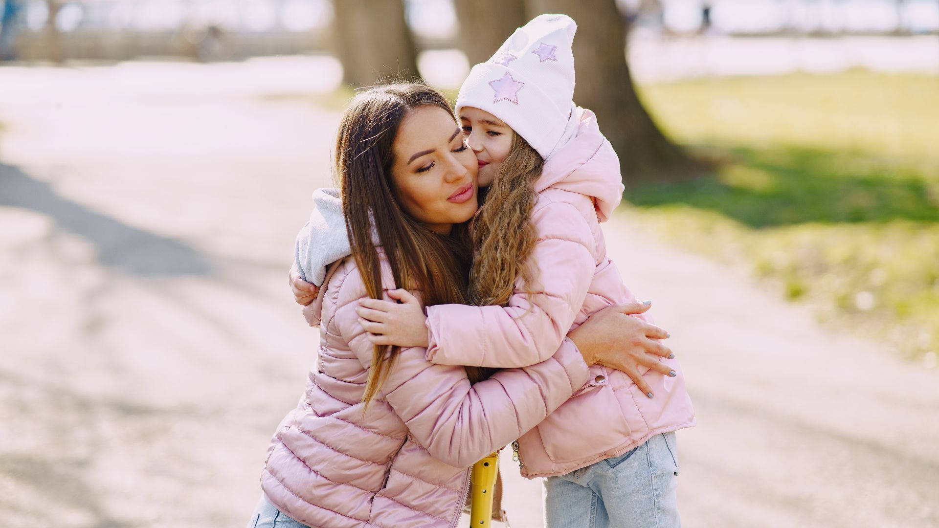 Mom 桌布 壁紙 — 一位母親和女兒穿著相配的粉紅色羽絨外套，在陽光明媚、有樹木和草地的公園裡溫馨擁抱。