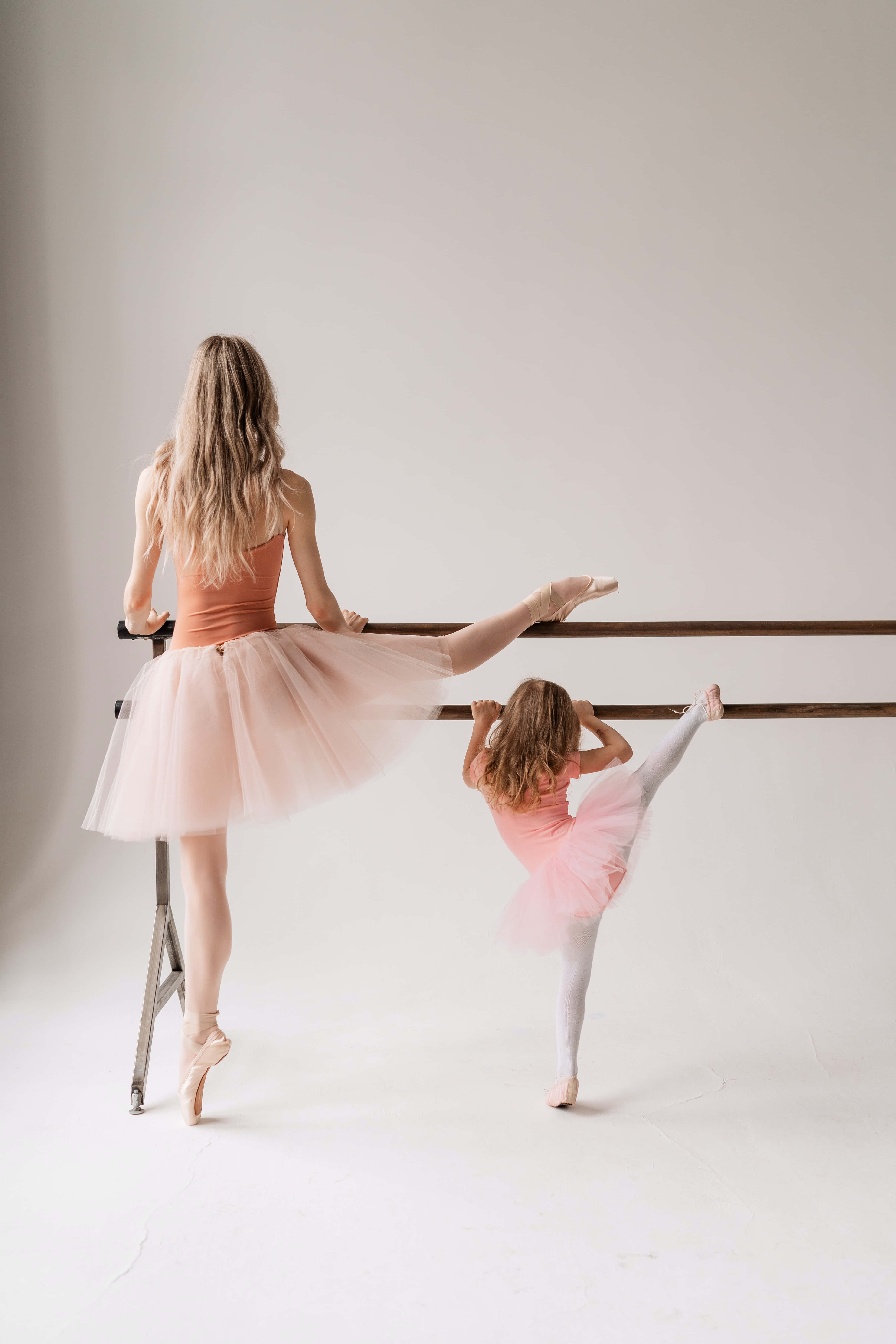 芭蕾 桌布 壁紙 — 一位母親和女兒穿著粉紅色芭蕾舞裙，在極簡白色工作室背景下優雅地在芭蕾舞把桿上伸展。