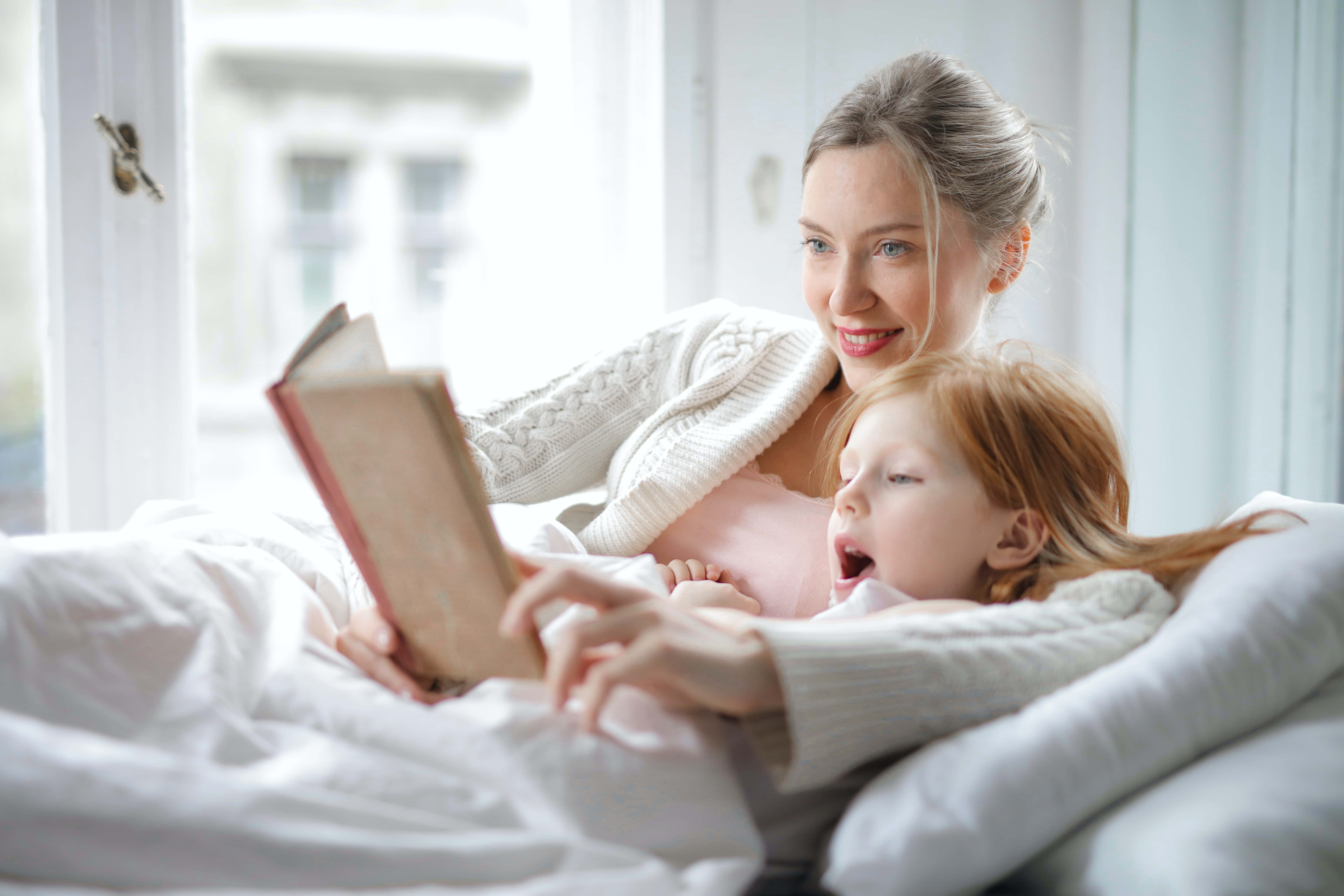 Mom 桌布 壁紙 — 一位母親和她年幼的女兒在床上蓋著白色被子依偎在一起，在柔和的光線下一起看書。