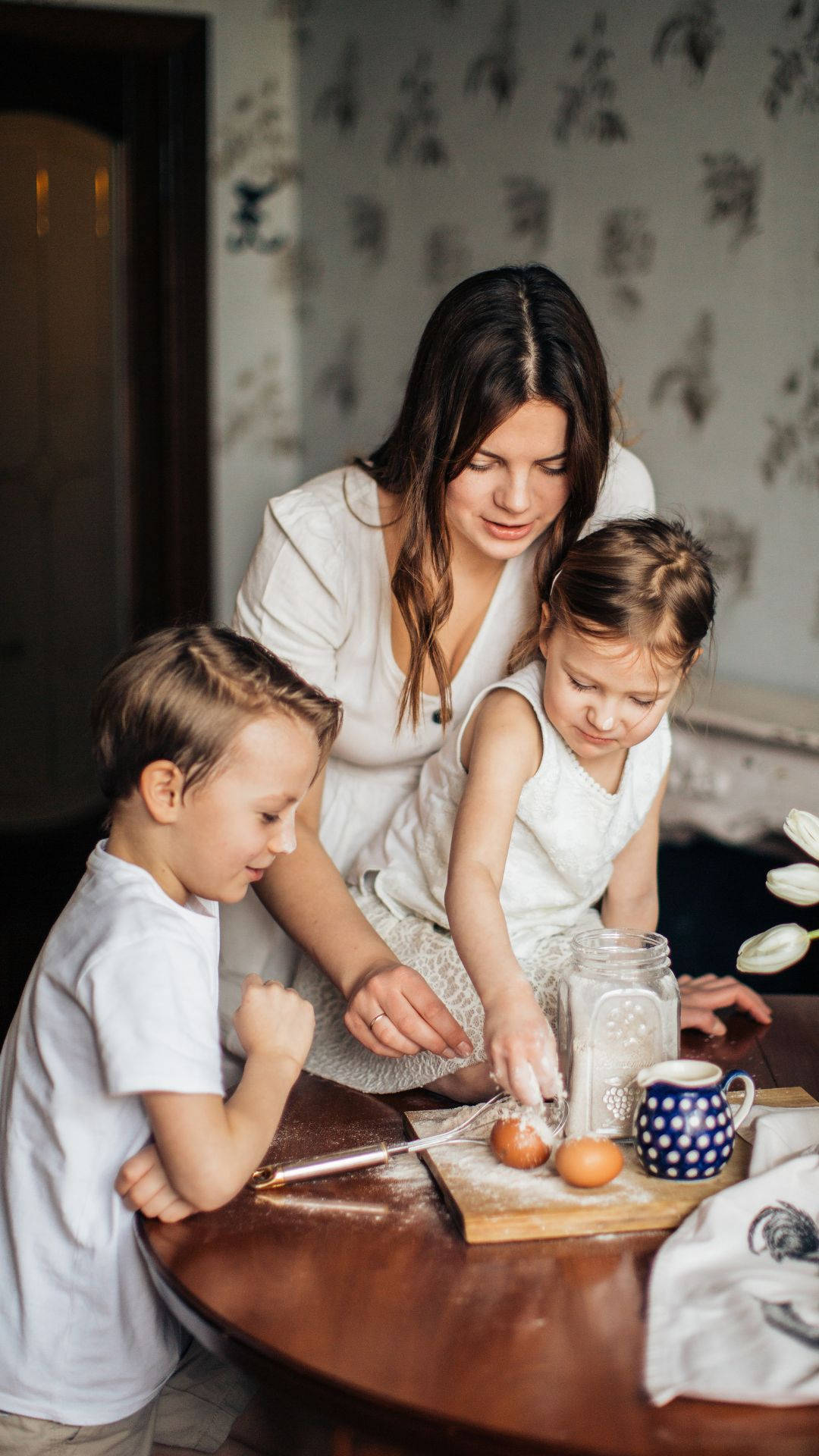 Mom 桌布 壁紙 — 一位母親和她的兩個孩子在溫暖照明的家庭廚房裡，在一張灑滿麵粉的木桌上一起烘焙。