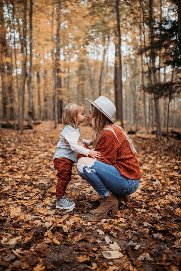 Family 桌布 壁紙 — 一位母親和孩子在陽光明媚的秋日森林中，被金色落葉覆蓋，分享一個溫柔的吻。