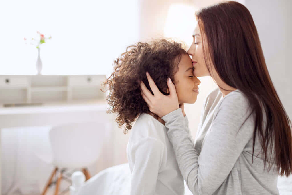 Mother Love 桌布 壁紙 — 一位母親在明亮、陽光充足的臥室中，溫柔地親吻女兒的額頭，房間充滿柔和自然光。