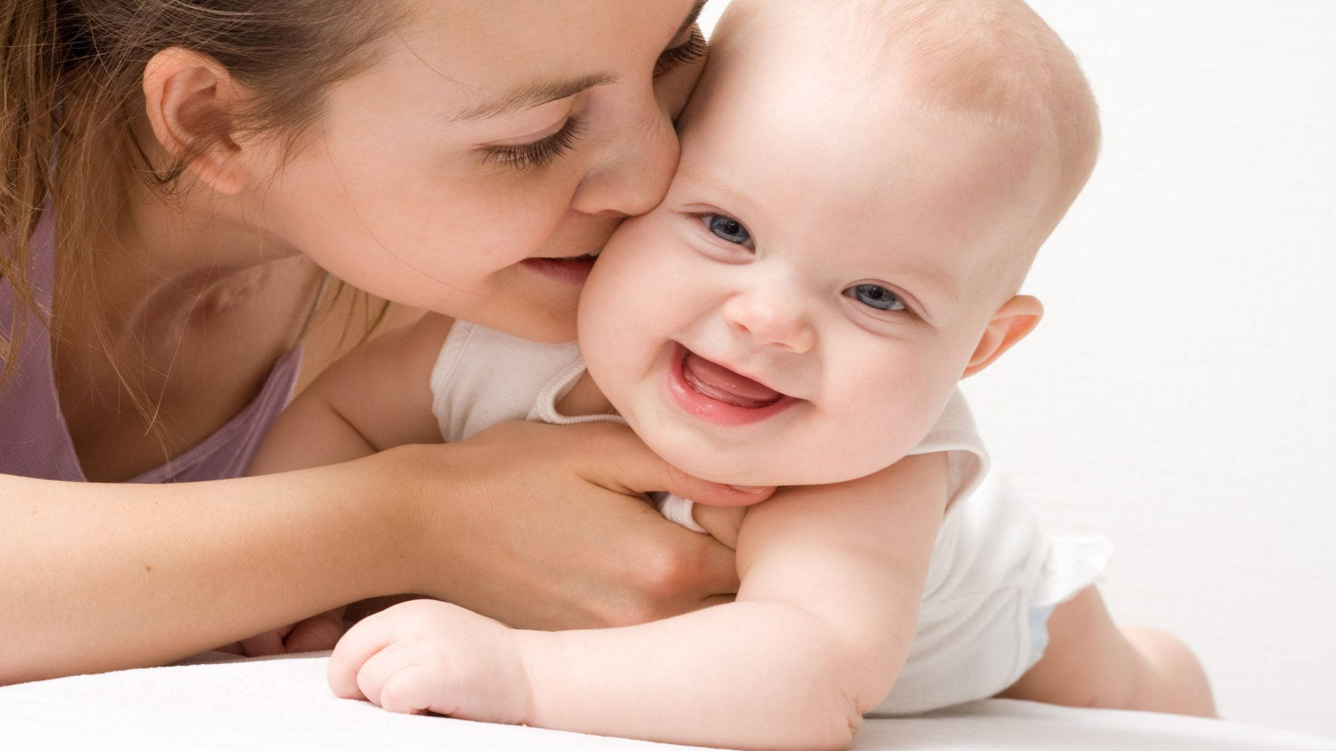 Funny Baby 桌布 壁紙 — 一位母親在柔和的白色背景下溫柔地親吻她微笑的嬰兒的臉頰。