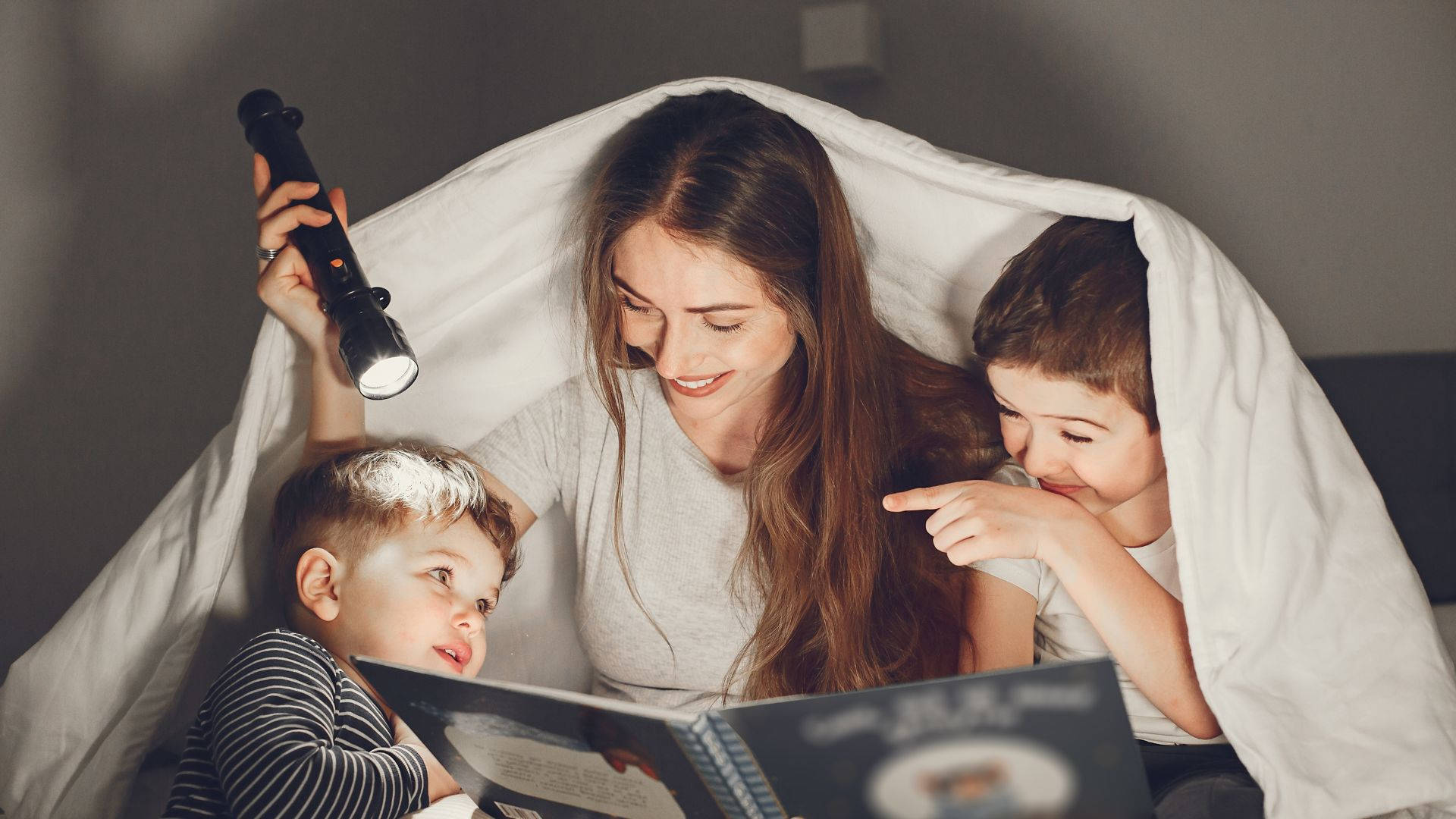 Mom 桌布 壁紙 — 一位母親在溫馨的夜晚環境中，用手電筒照亮白色毯子，為兩個孩子讀書。