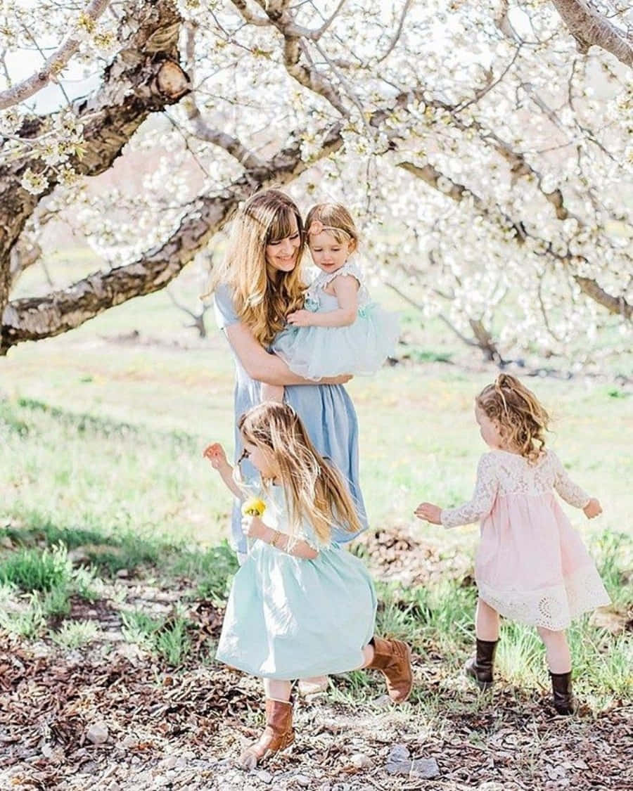 Spring Family 桌布 壁紙 — 一位母親在盛開的櫻花樹下擁抱她的幼兒，三個年幼的女兒在斑駁的陽光下玩耍。