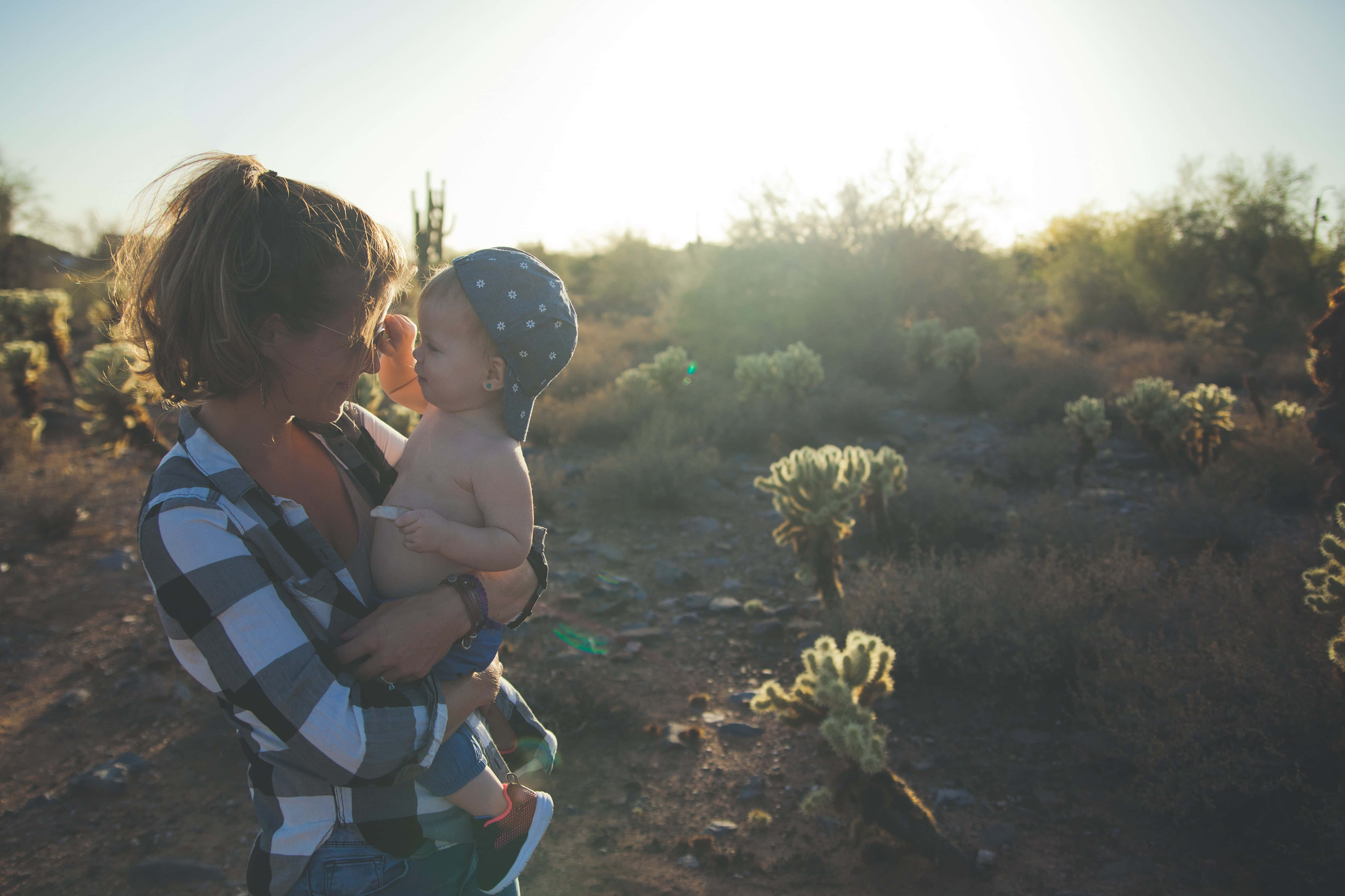Mom 桌布 壁紙 — 一位母親在陽光明媚、點綴著仙人掌和柔和金色光線的沙漠景觀中，溫柔地抱著她的嬰兒。