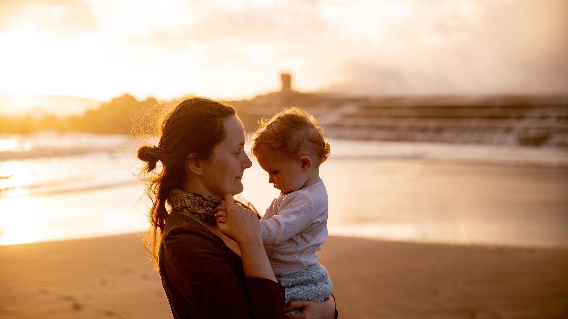 Mom 桌布 壁紙 — 一位母親在陽光明媚的海灘上溫柔地抱著她的嬰兒，金色的海浪在日落時分翻滾而來。