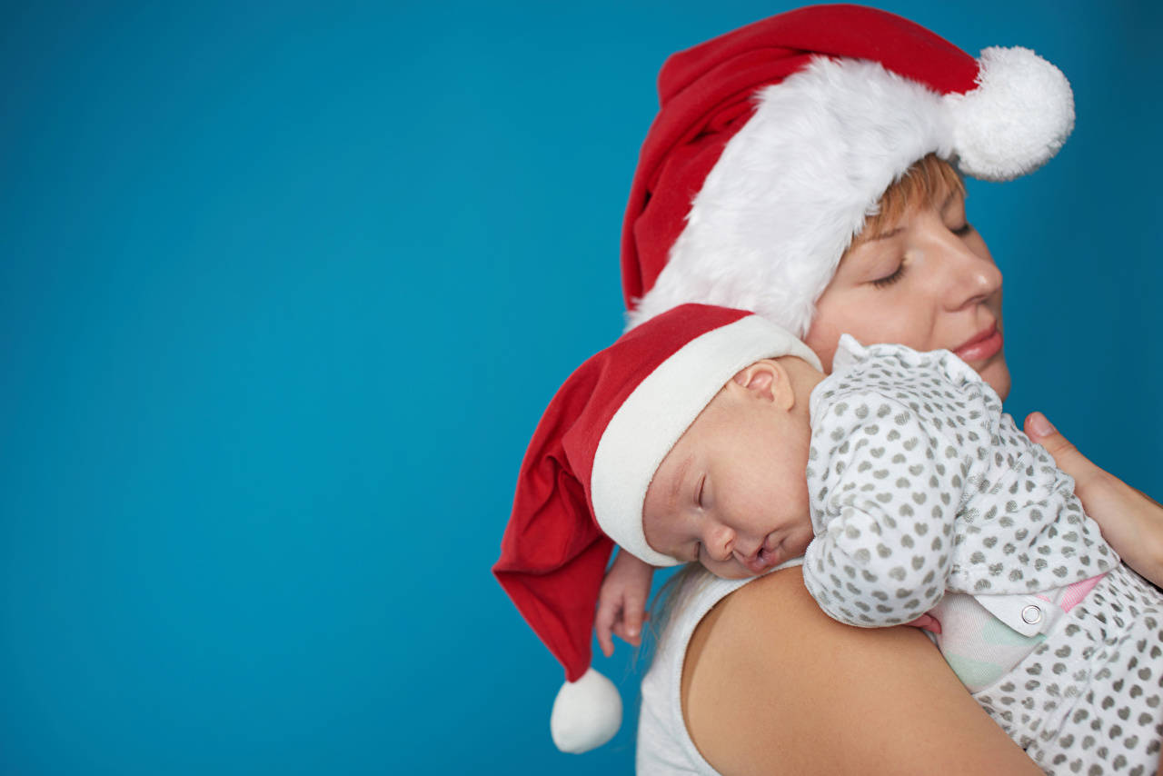 Santa 桌布 壁紙 — 一位母親懷抱著一個熟睡的嬰兒，兩人都戴著喜慶的紅白聖誕帽，背景是純藍色。