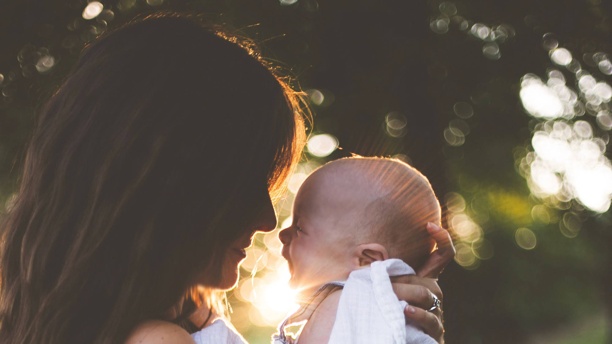 Mother And Baby 桌布 壁紙 — 一位母親溫柔地抱著她的寶寶，金色的陽光穿過樹木，在他們身後營造出溫暖發光的散景效果。