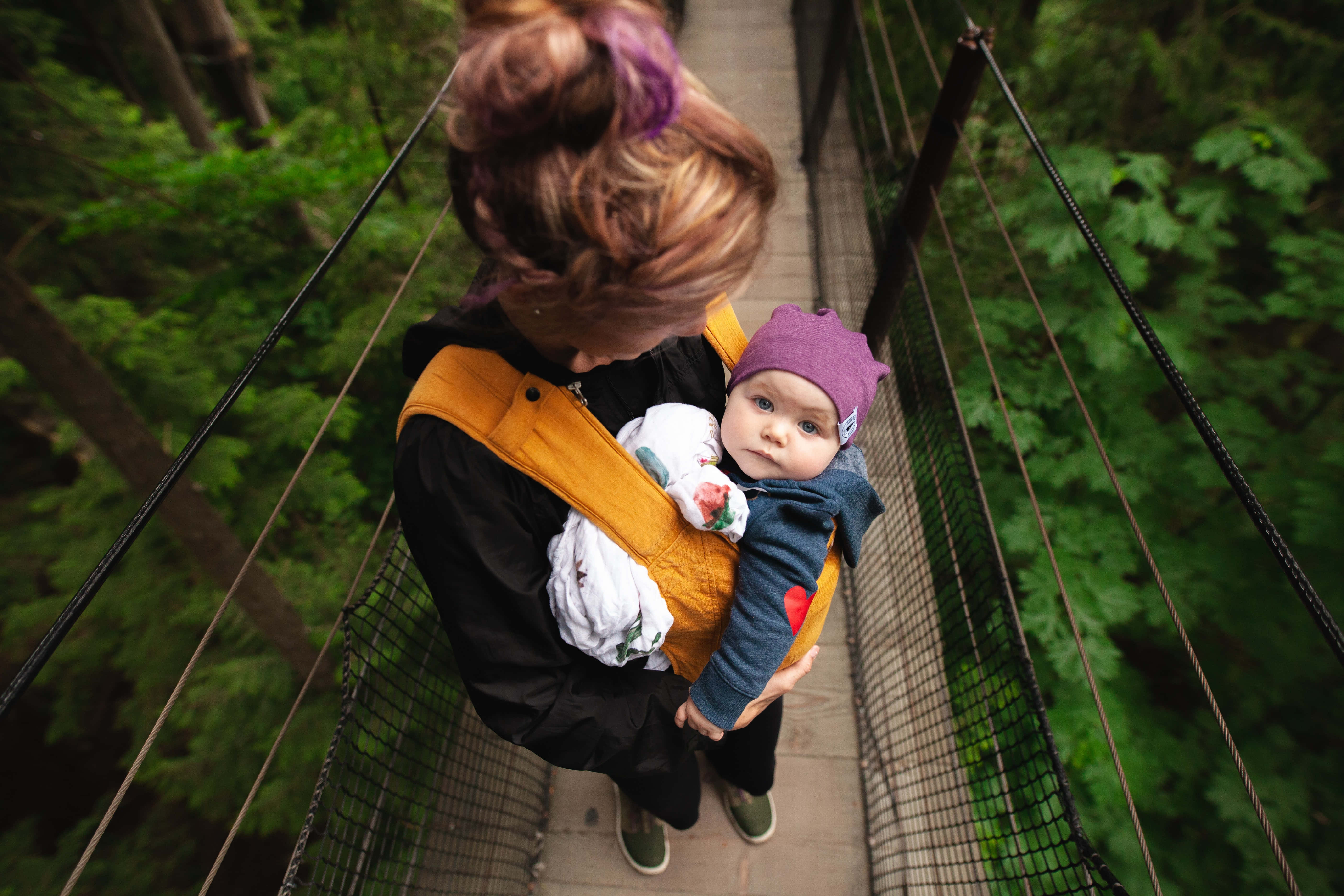 Mom 桌布 壁紙 — 一位母親用嬰兒揹帶揹著寶寶，走過一座被翠綠樹木環繞的森林吊橋。