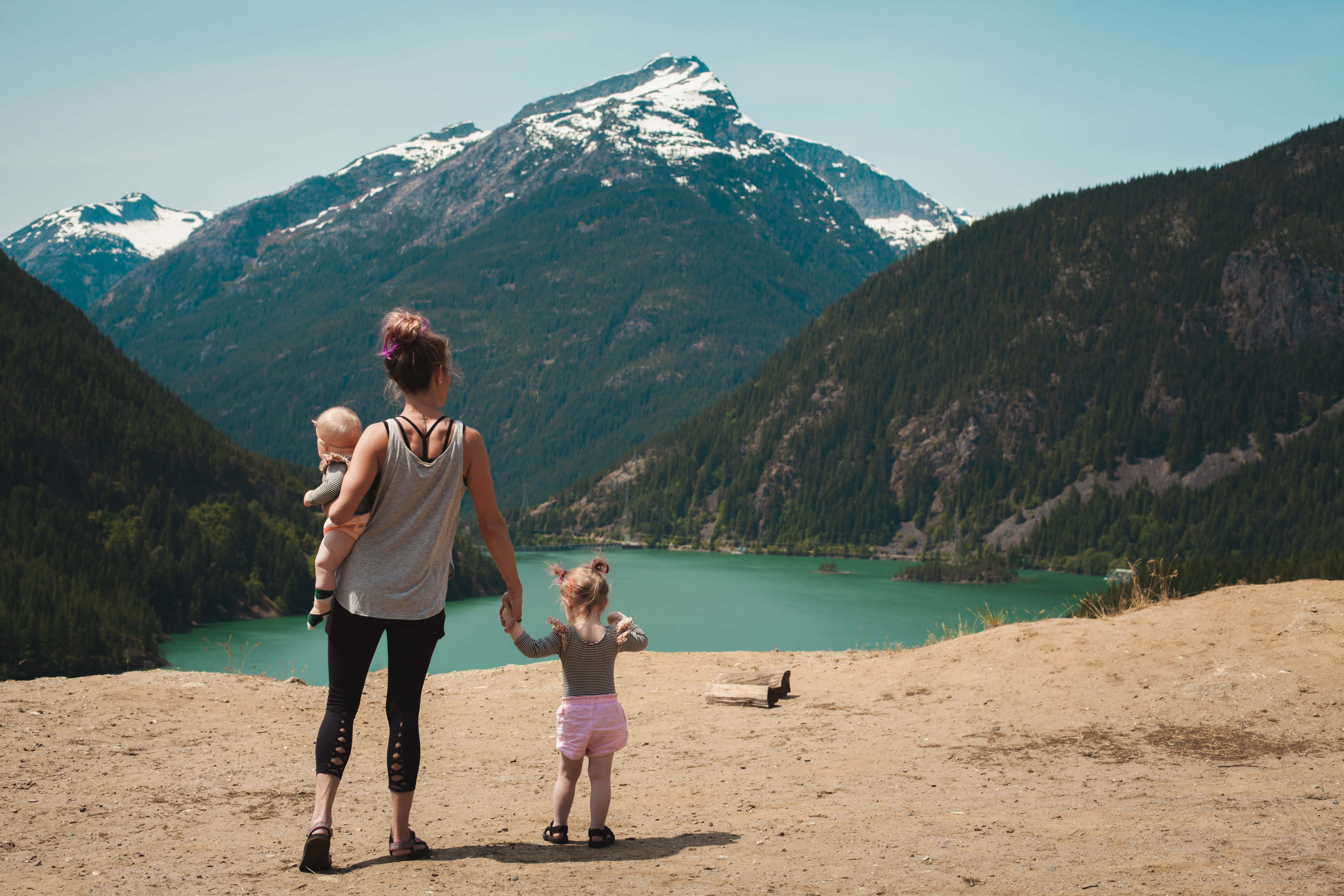 Mom 桌布 壁紙 — 一位母親用揹帶揹著嬰兒，並牽著一個幼兒的手，凝視著一個被雪山環繞的碧綠湖泊。