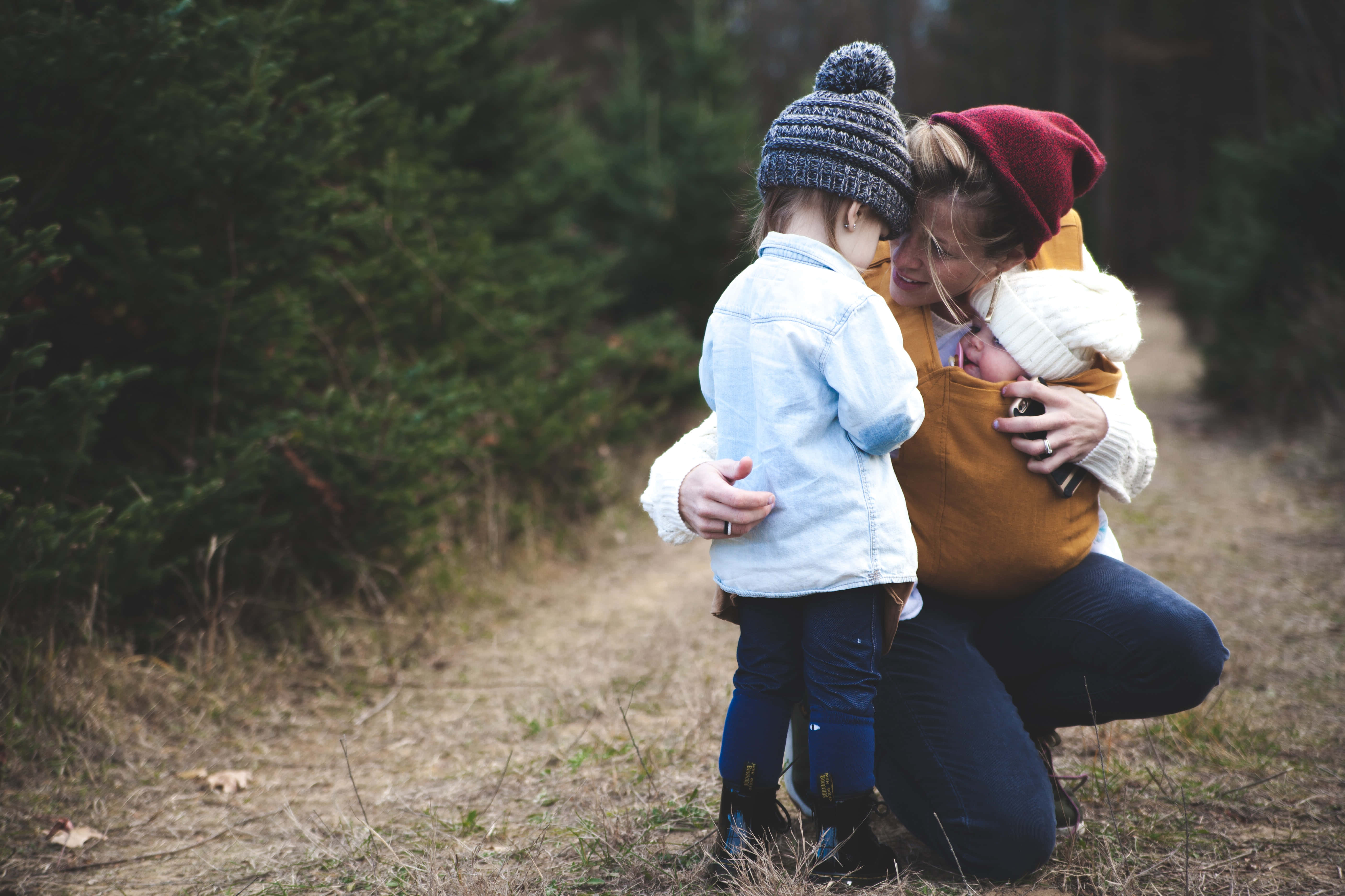 Mom 桌布 壁紙 — 一位母親跪在森林裡，用揹帶揹著嬰兒，同時擁抱著一個戴著針織帽的年長孩子。