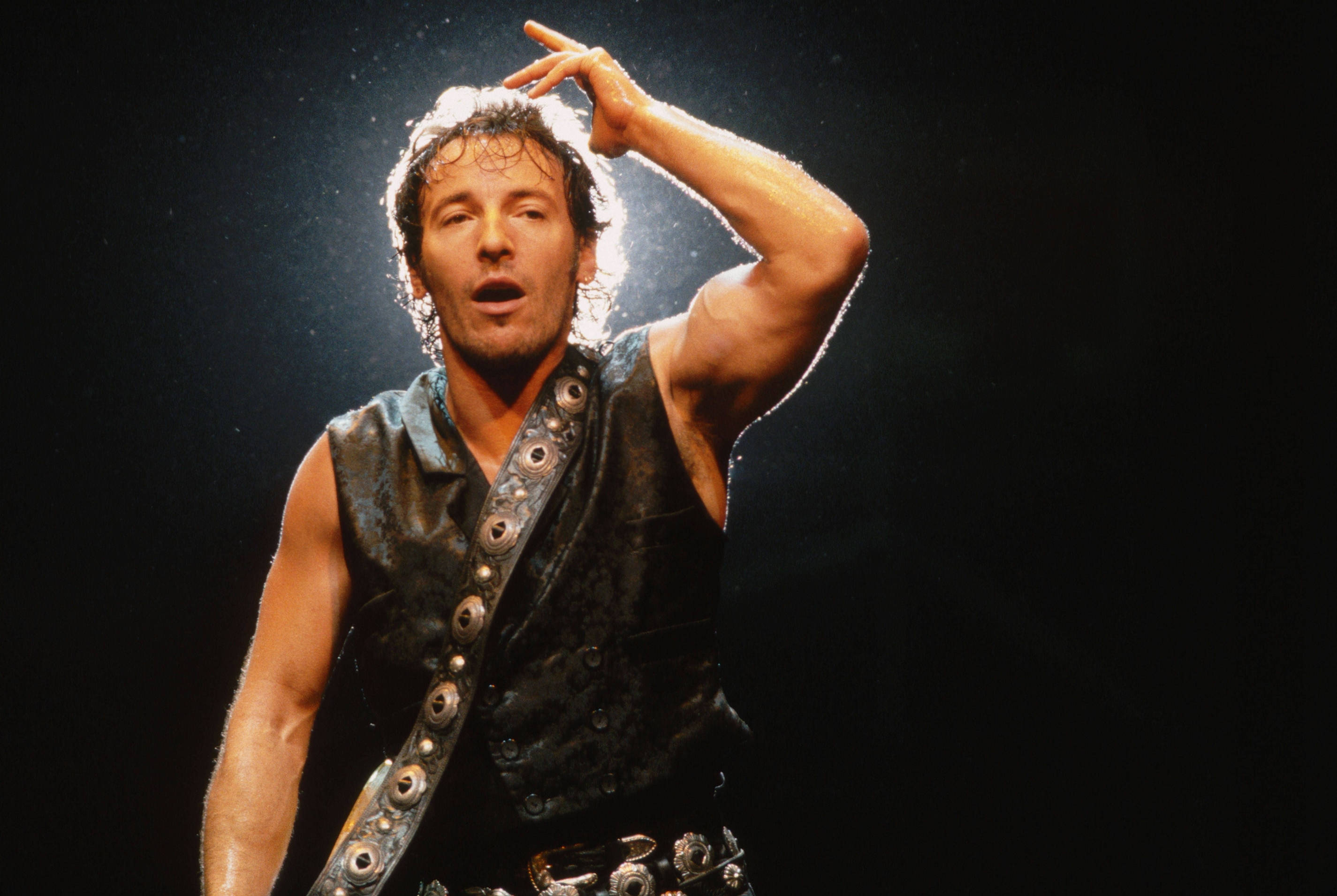 Bruce Springsteen 桌布 壁紙 — 一位汗流浹背的男性表演者穿著鉚釘背心，在黑色背景的明亮舞台燈光下熱情歌唱。