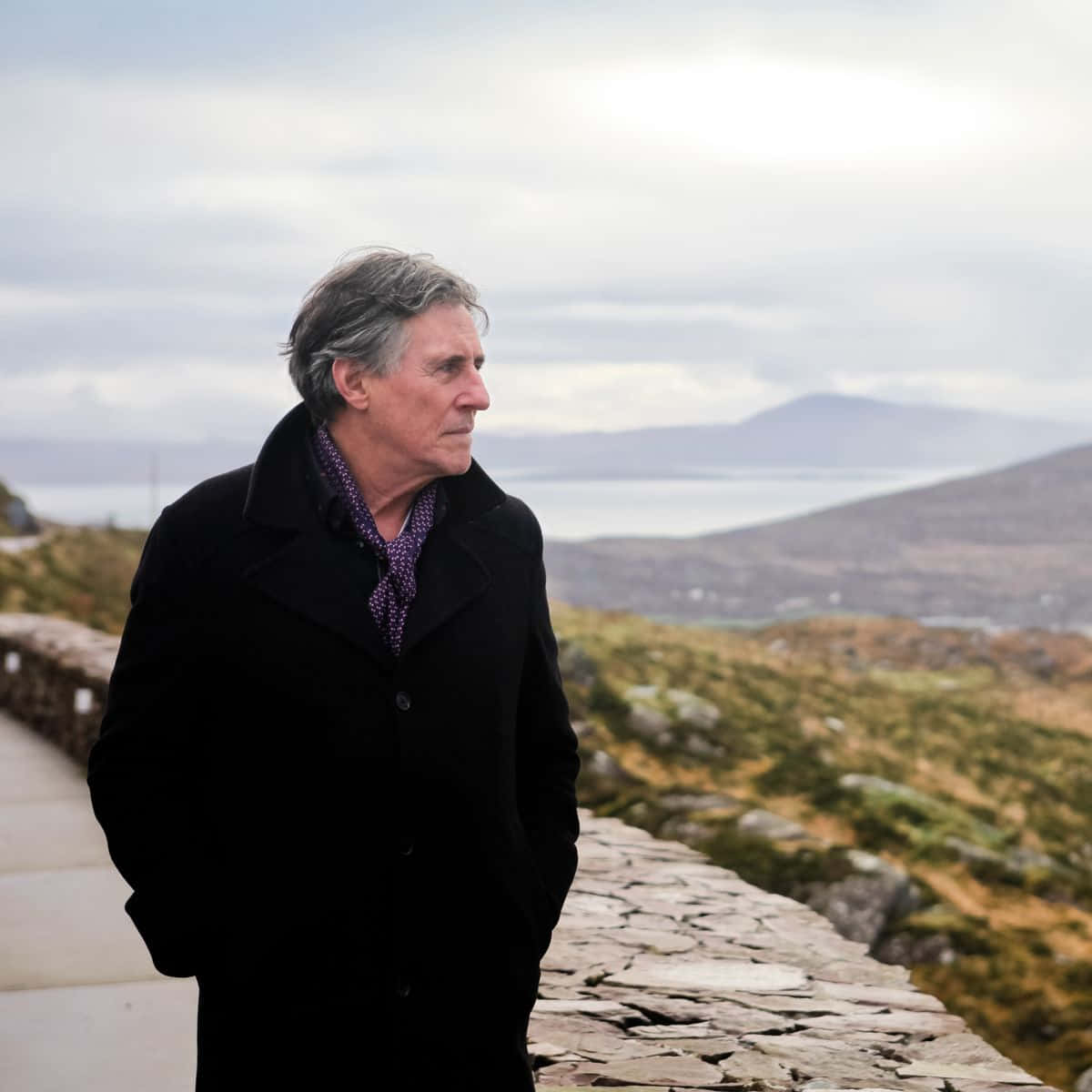 Gabriel Byrne 桌布 壁紙 — 一位沉思的老年男子身穿黑色外套，站在一條石徑上，俯瞰著霧氣瀰漫的沿海風景，遠處有山丘。