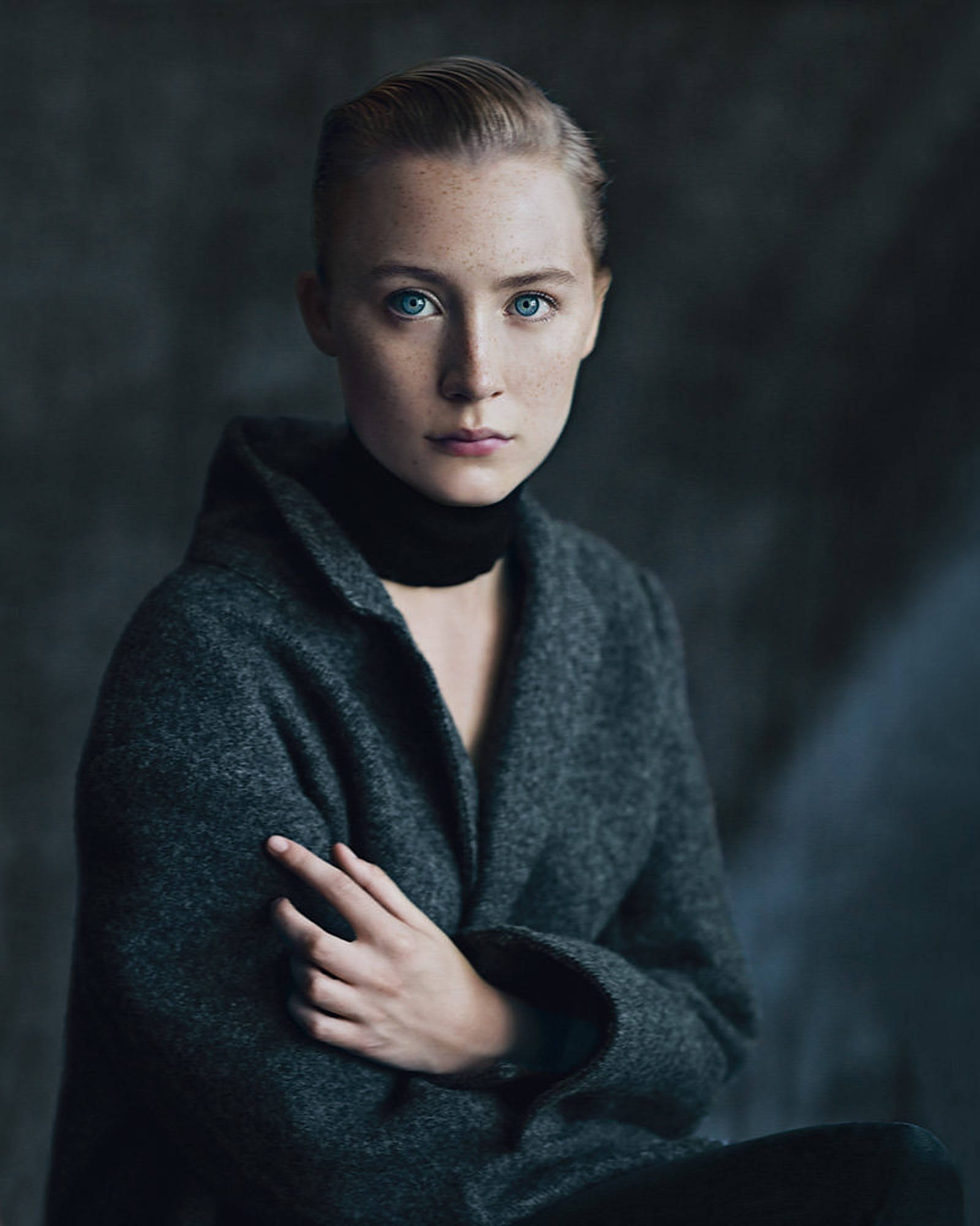 Saoirse Ronan 桌布 壁紙 — 一位沉穩的女性，擁有引人注目的藍色眼睛和雀斑，直視鏡頭，身穿深灰色外套內搭黑色高領毛衣，背景為陰鬱的紋理背景。