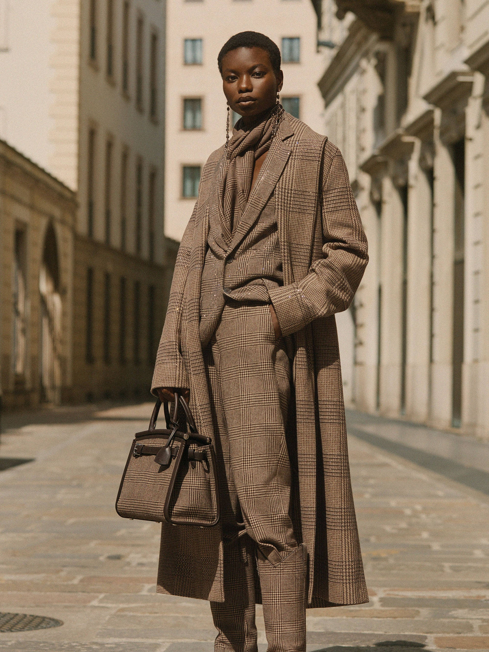 ralph lauren 桌布 壁紙 — 一位沉穩的女性身穿棕色格紋外套，自信地站在陽光普照、擁有歷史建築的歐洲街道上。
