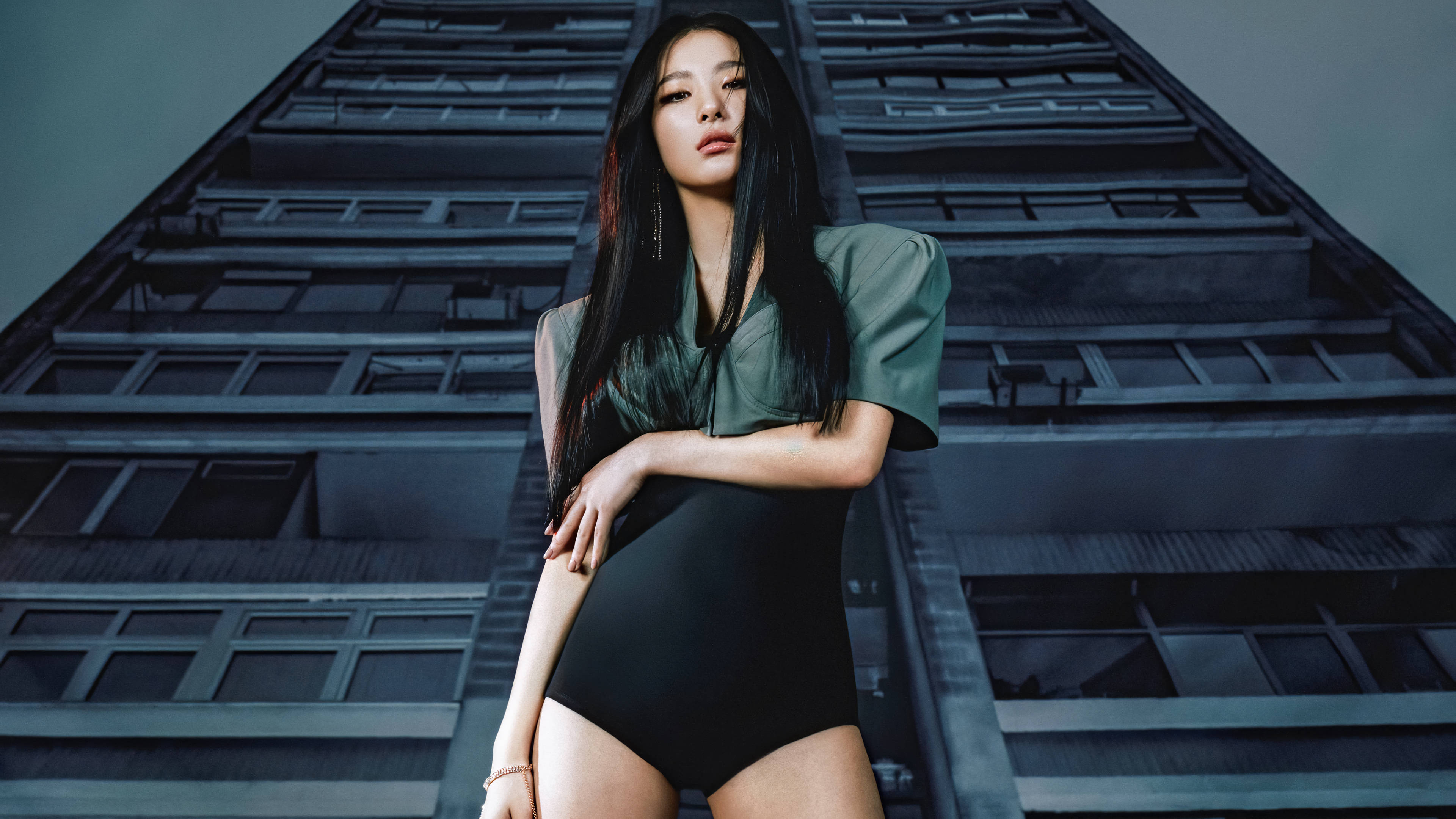 Velvet 桌布 壁紙 — 一位沉穩的女性身穿綠色上衣和黑色連體衣，在冷藍色燈光下自信地站在高聳的城市建築前。