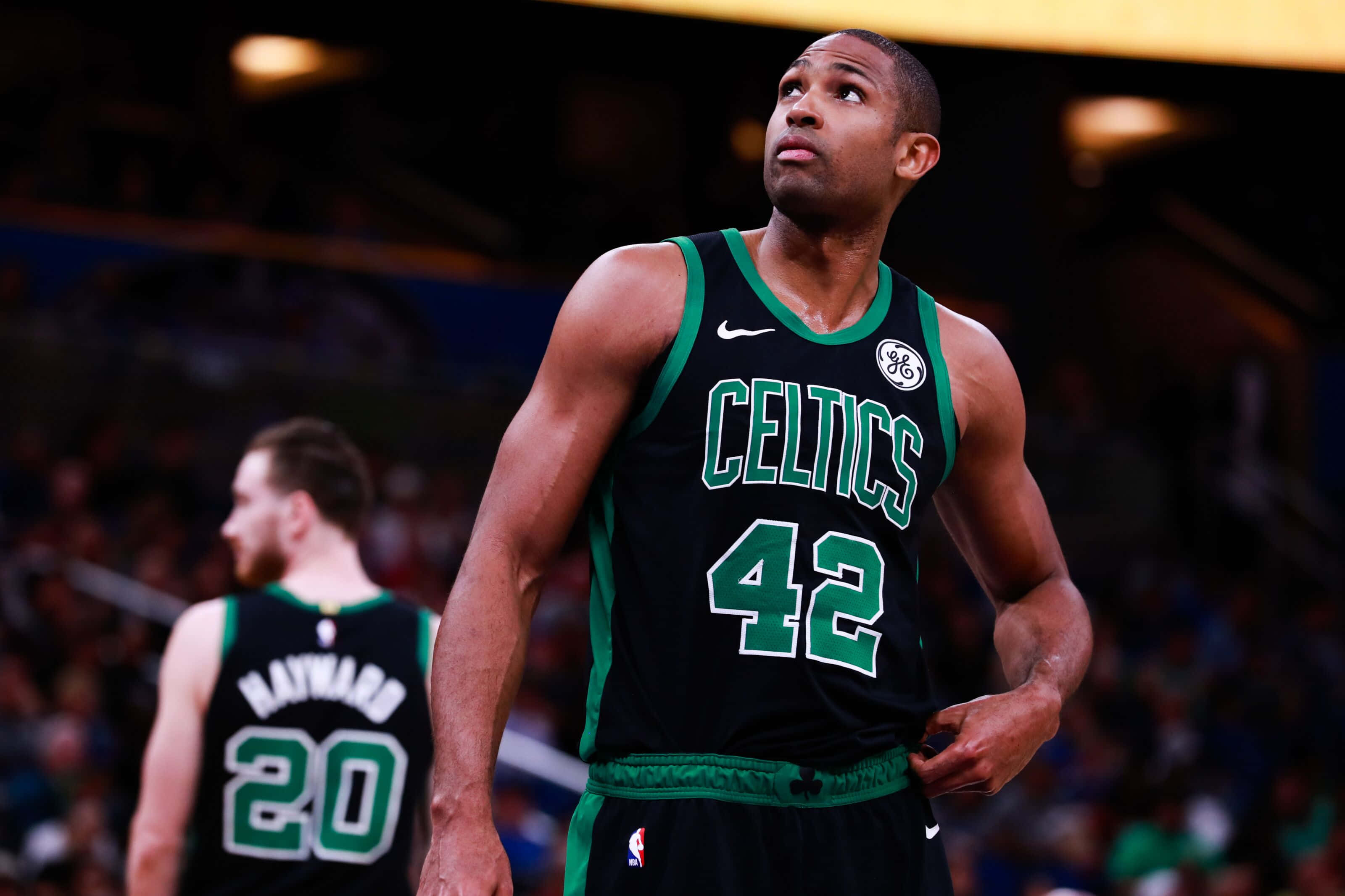 Al Horford 桌布 壁紙 — 一位波士頓塞爾提克籃球運動員身穿黑色和綠色球衣（號碼42）在場館燈光下比賽時向上看。