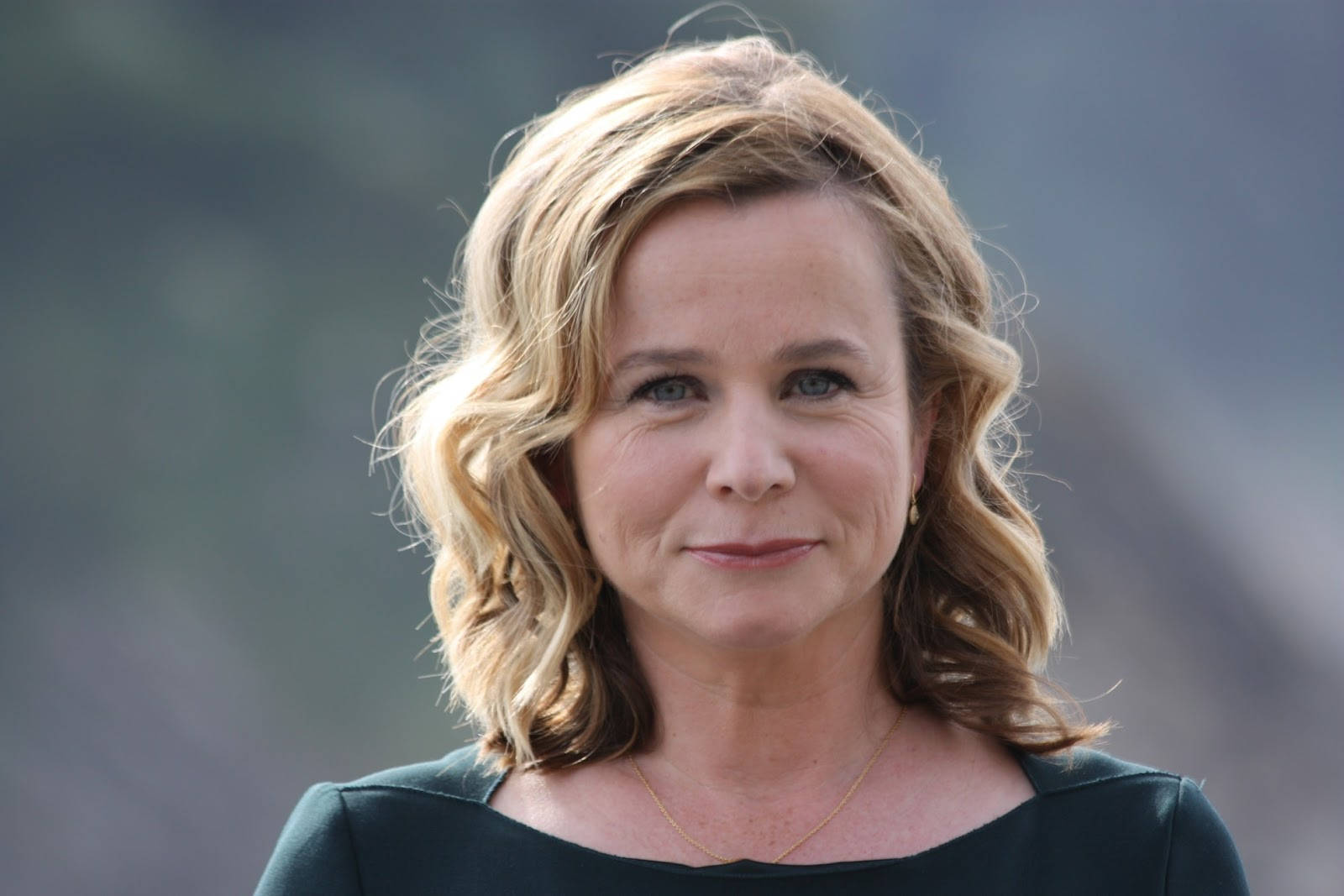 女演員 桌布 壁紙 — 一位波浪金髮、溫柔微笑的女性特寫肖像，身穿深綠色上衣。