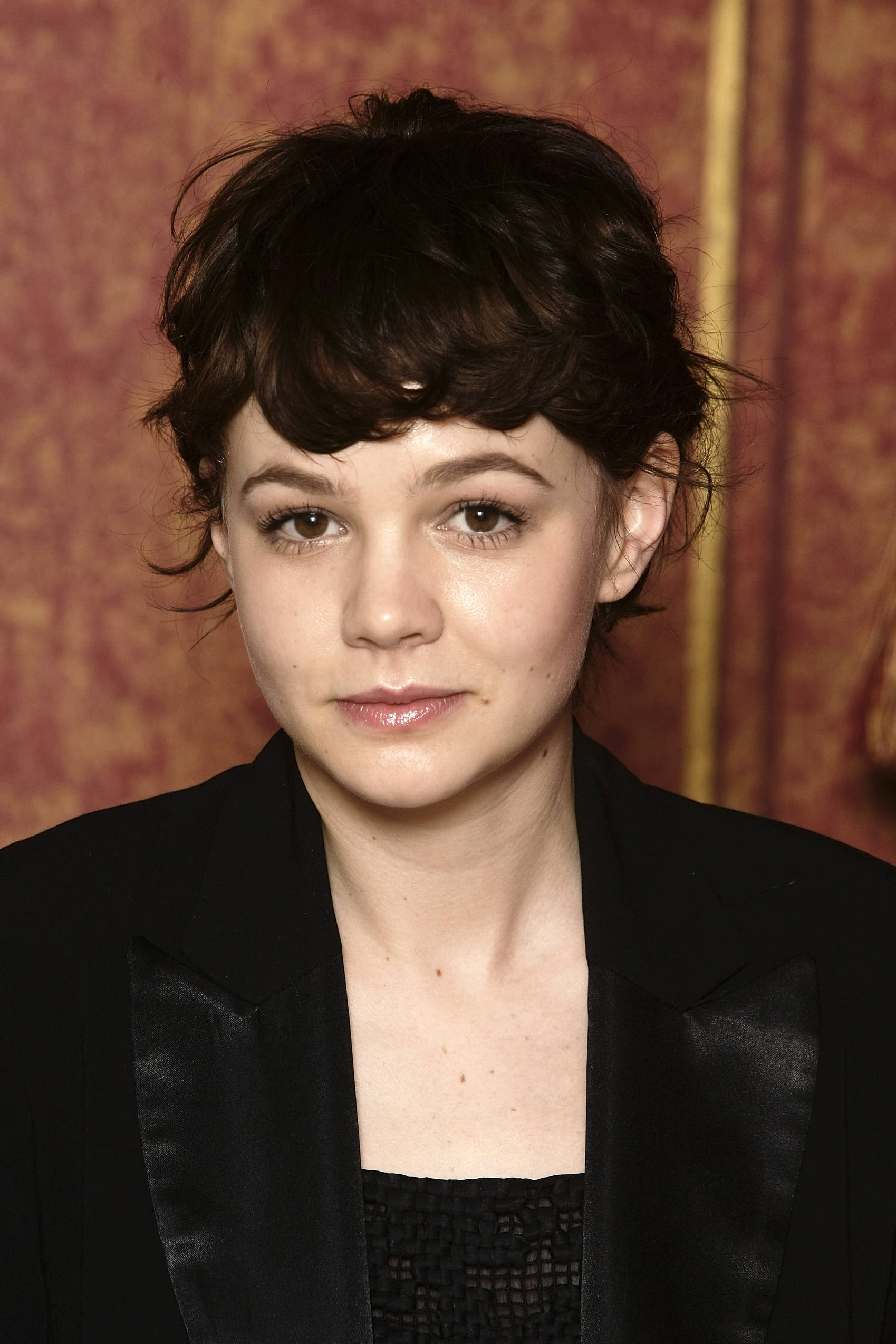 Carey Mulligan 桌布 壁紙 — 一位深色亂髮、身穿黑色西裝外套的女性，在豐富的紅色圖案背景下的特寫肖像。