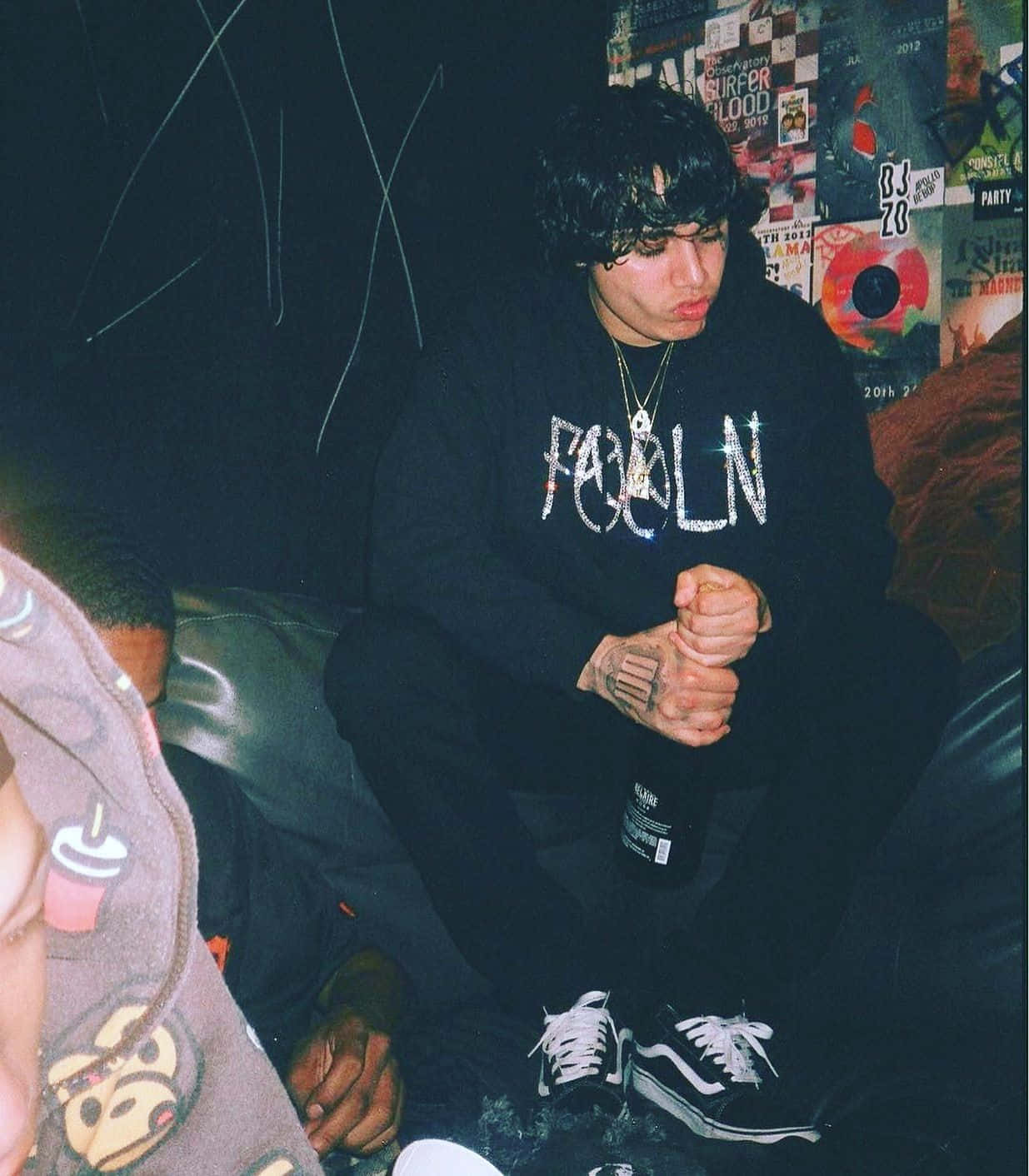 Shoreline Mafia 桌布 壁紙 — 一位深色捲髮的年輕男子坐在光線昏暗、貼滿海報和塗鴉的沙發上。