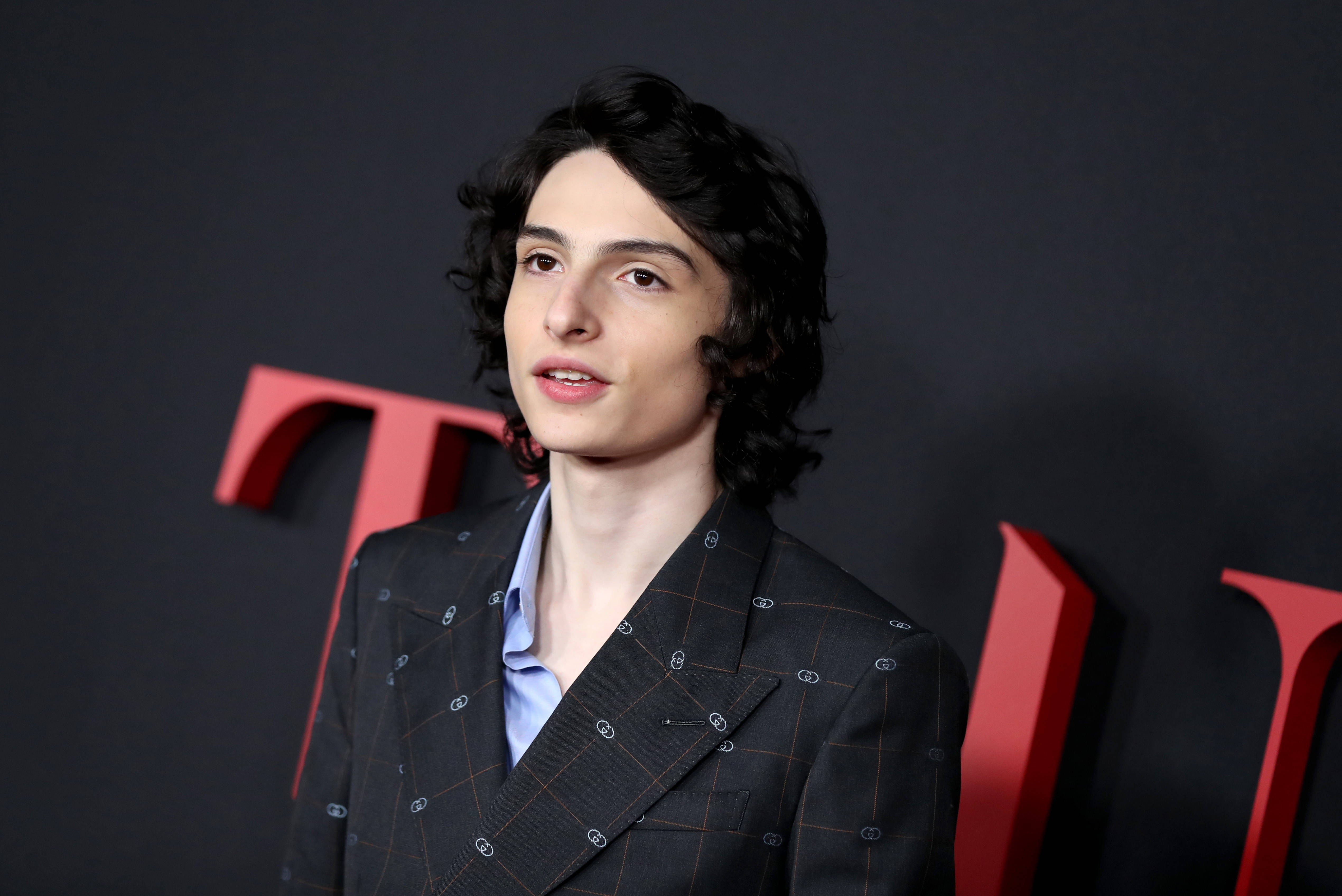 Celebrity 桌布 壁紙 — 一位深色捲髮的年輕男子穿著圖案西裝，背景為深色並帶有紅色字樣。