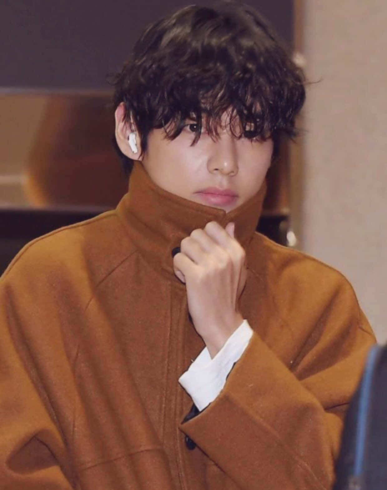 Taehyung Cute 桌布 壁紙 — 一位深色捲髮的年輕男子穿著棕色外套，戴著白色耳塞，在室內整理衣領。