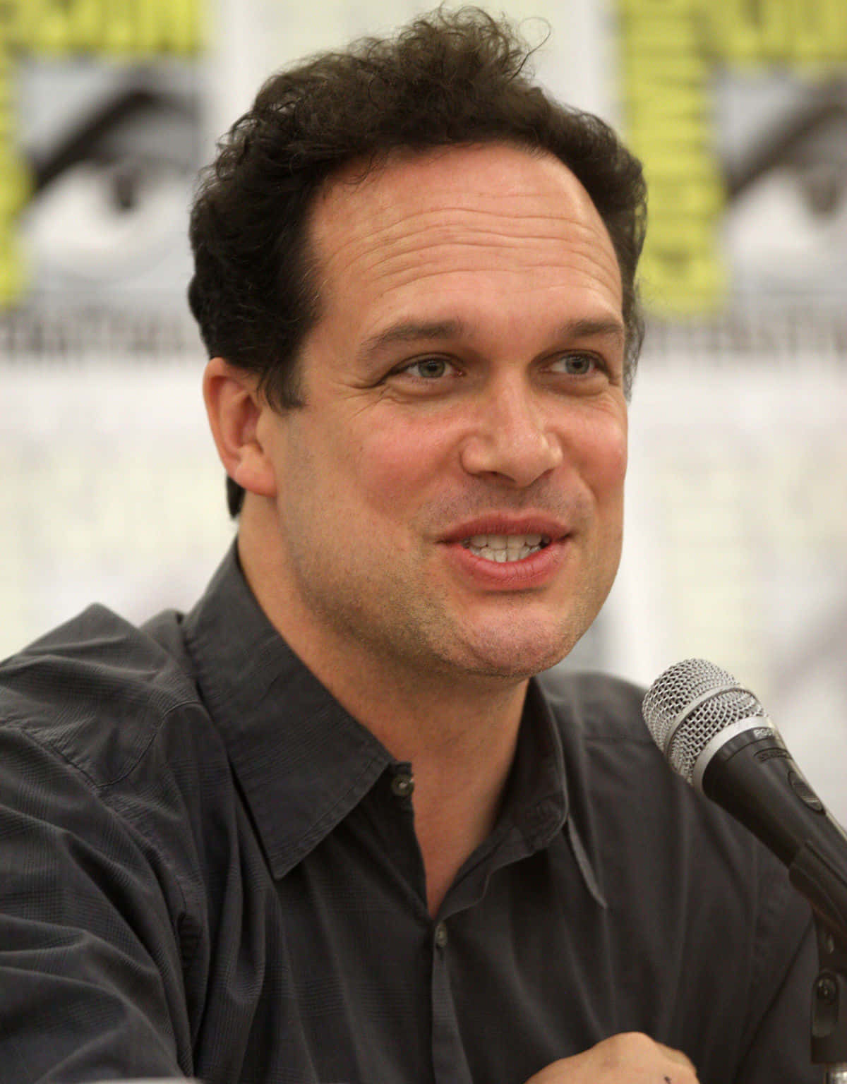Diedrich Bader 桌布 壁紙 — 一位深色捲髮的男子在會議小組中對著麥克風微笑說話。