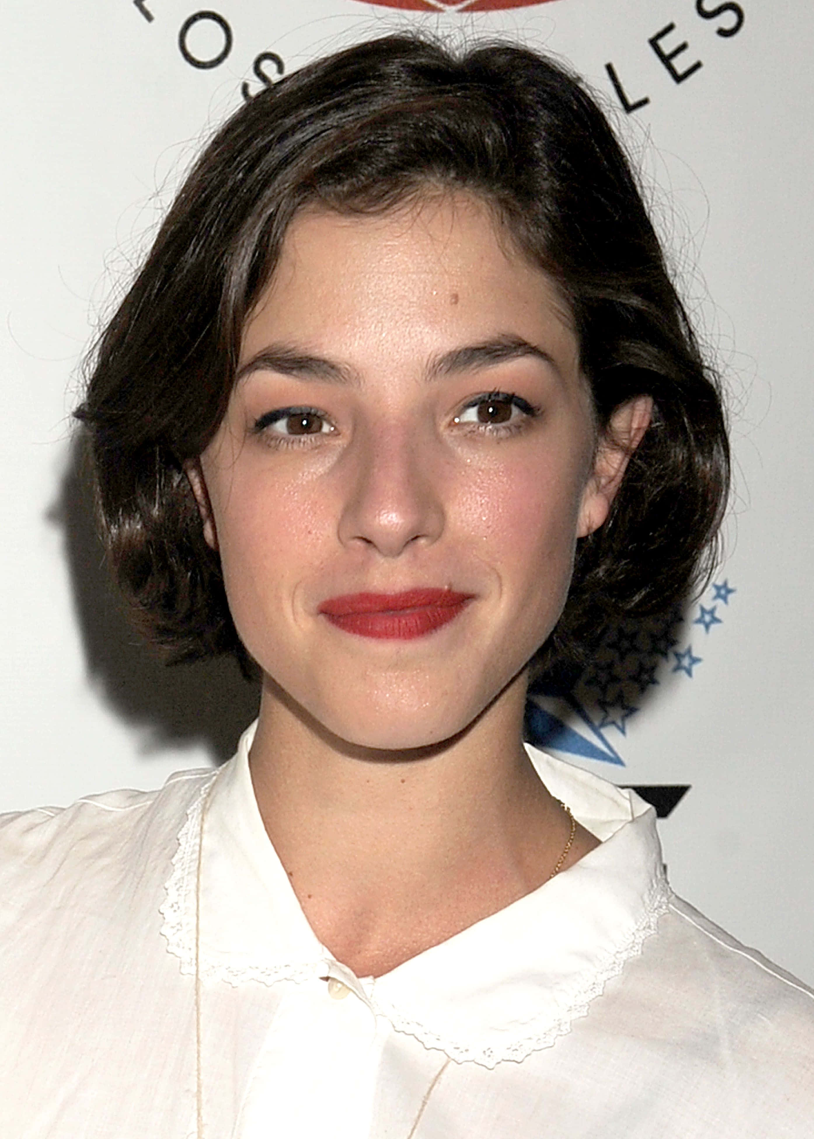 Olivia Thirlby 桌布 壁紙 — 一位深色波浪髮、塗紅色唇膏、身穿白色襯衫的女子的特寫肖像，背景有部分文字和星星。
