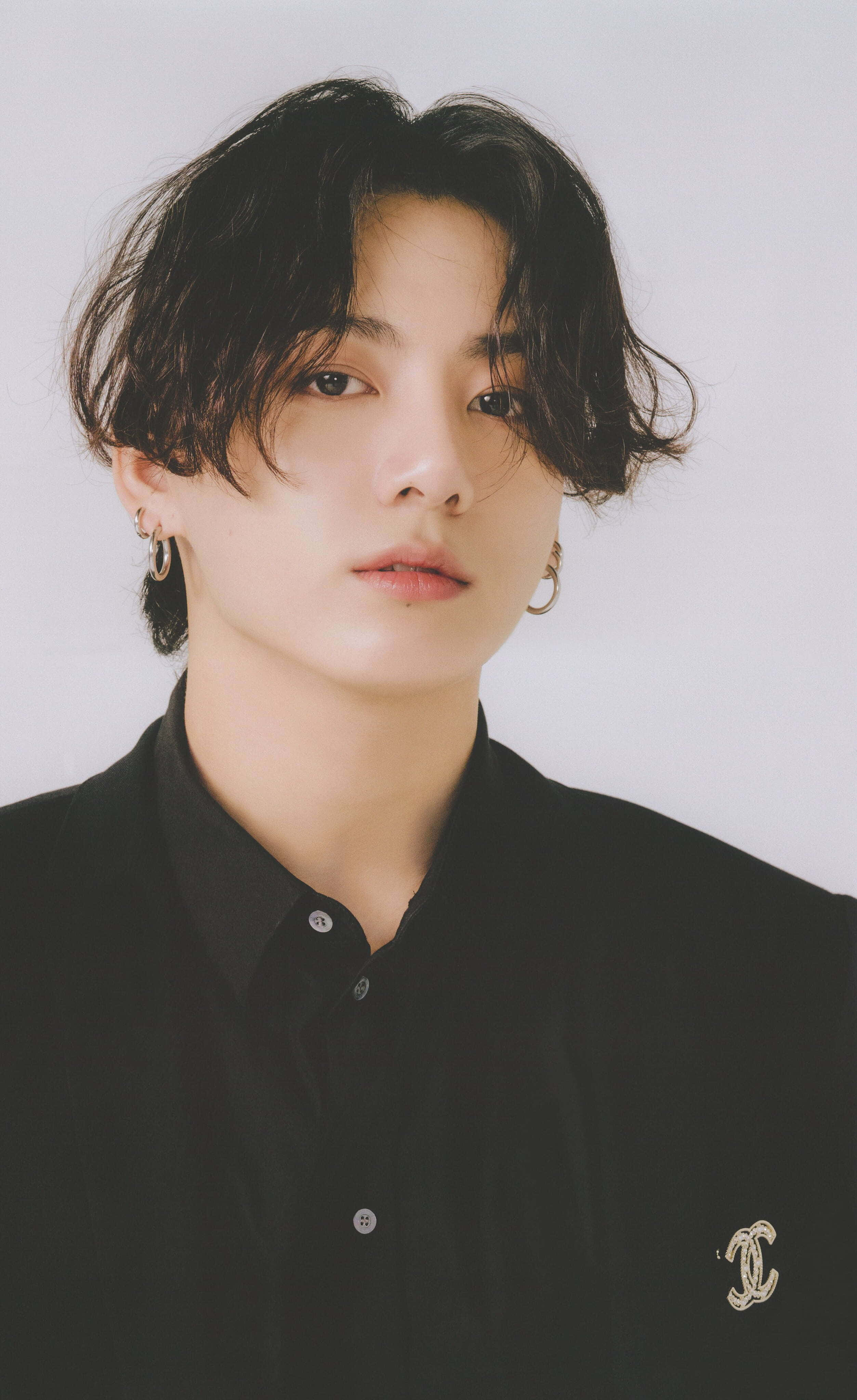 Jungkook 桌布 壁紙 — 一位深色波浪髮、戴金色圓環耳環的年輕男子，身穿黑色鈕扣襯衫，別著Chanel標誌胸針，背景為純白色。