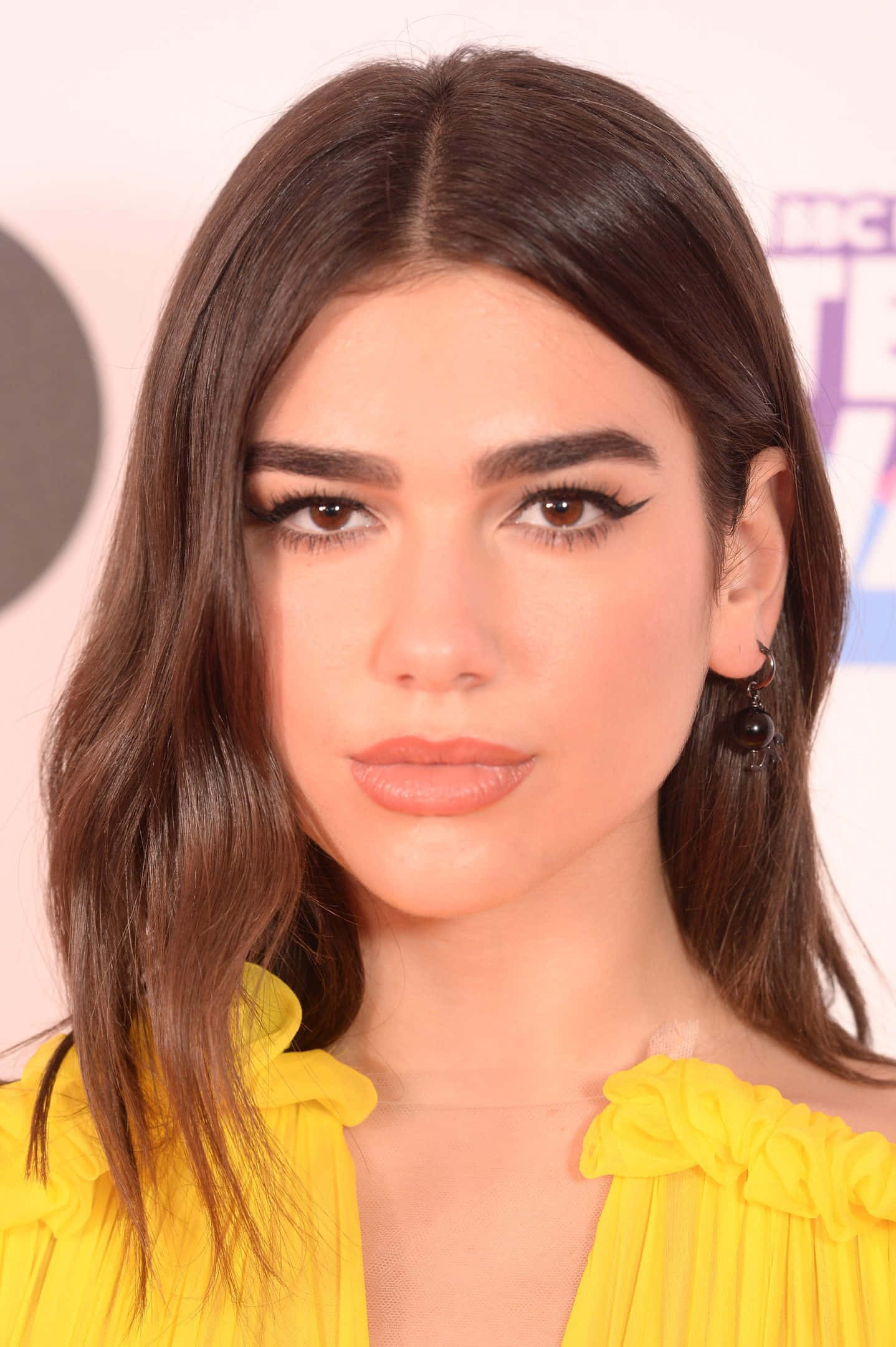 Dua Lipa 桌布 壁紙 — 一位深色波浪髮、濃黑眼線的女性穿著亮黃色荷葉邊上衣的頭像特寫。
