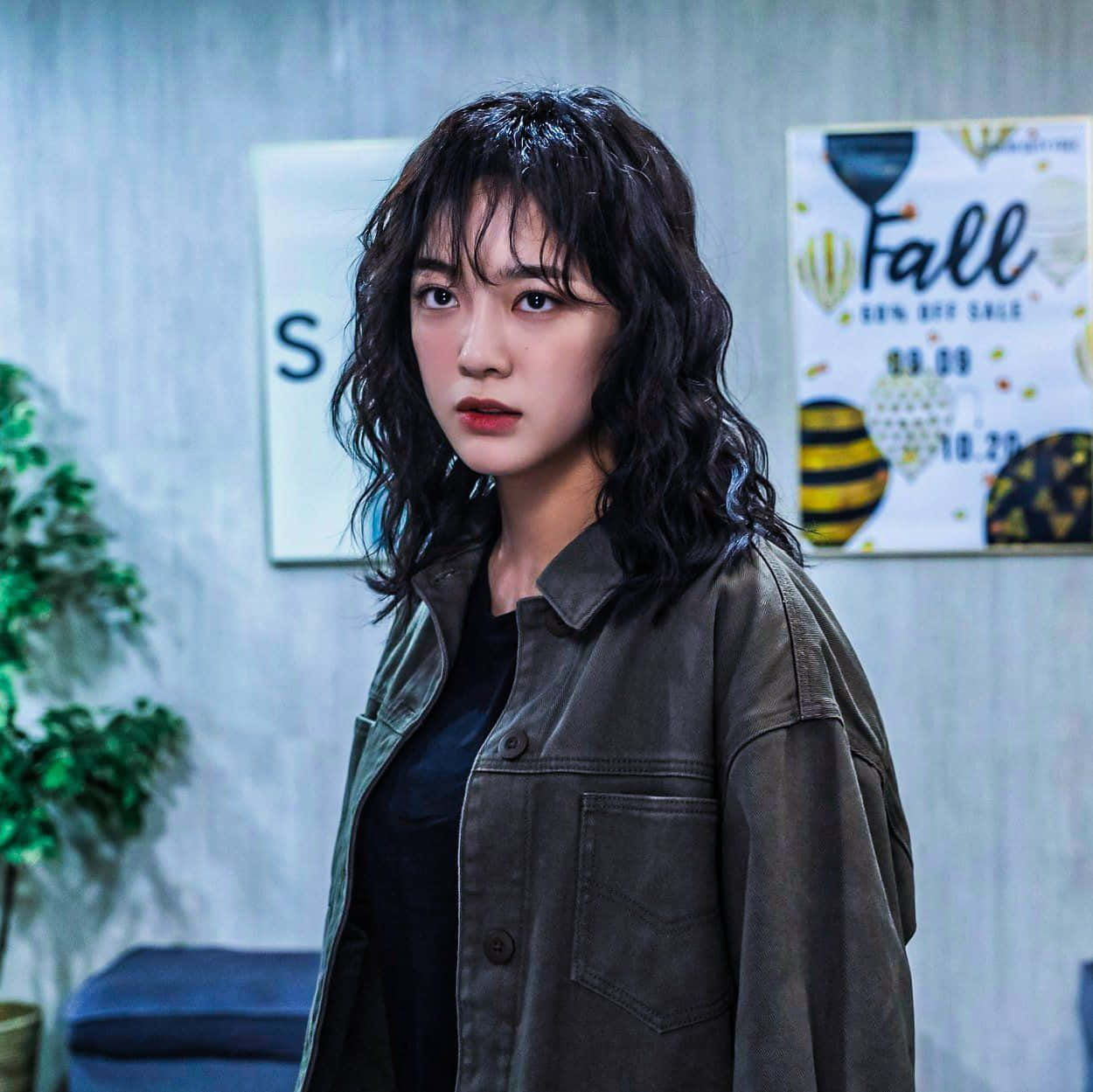 korean 桌布 壁紙 — 一位深色波浪髮、紅唇的年輕女子在一個現代房間裡，房間裡有秋季主題海報和盆栽植物，她專注地凝視著。