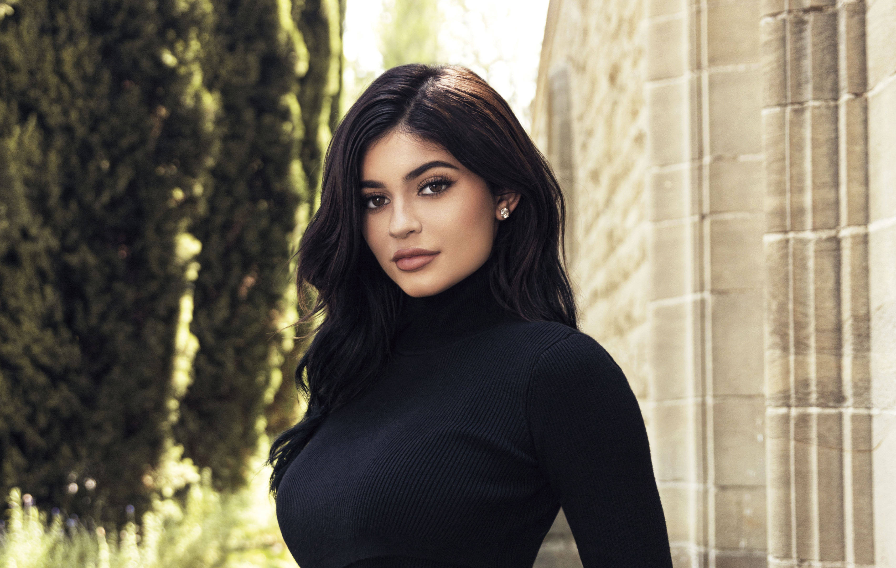 Kylie 桌布 壁紙 — 一位深色波浪髮、身穿黑色高領毛衣的女性站在石柱旁，身後有綠樹。