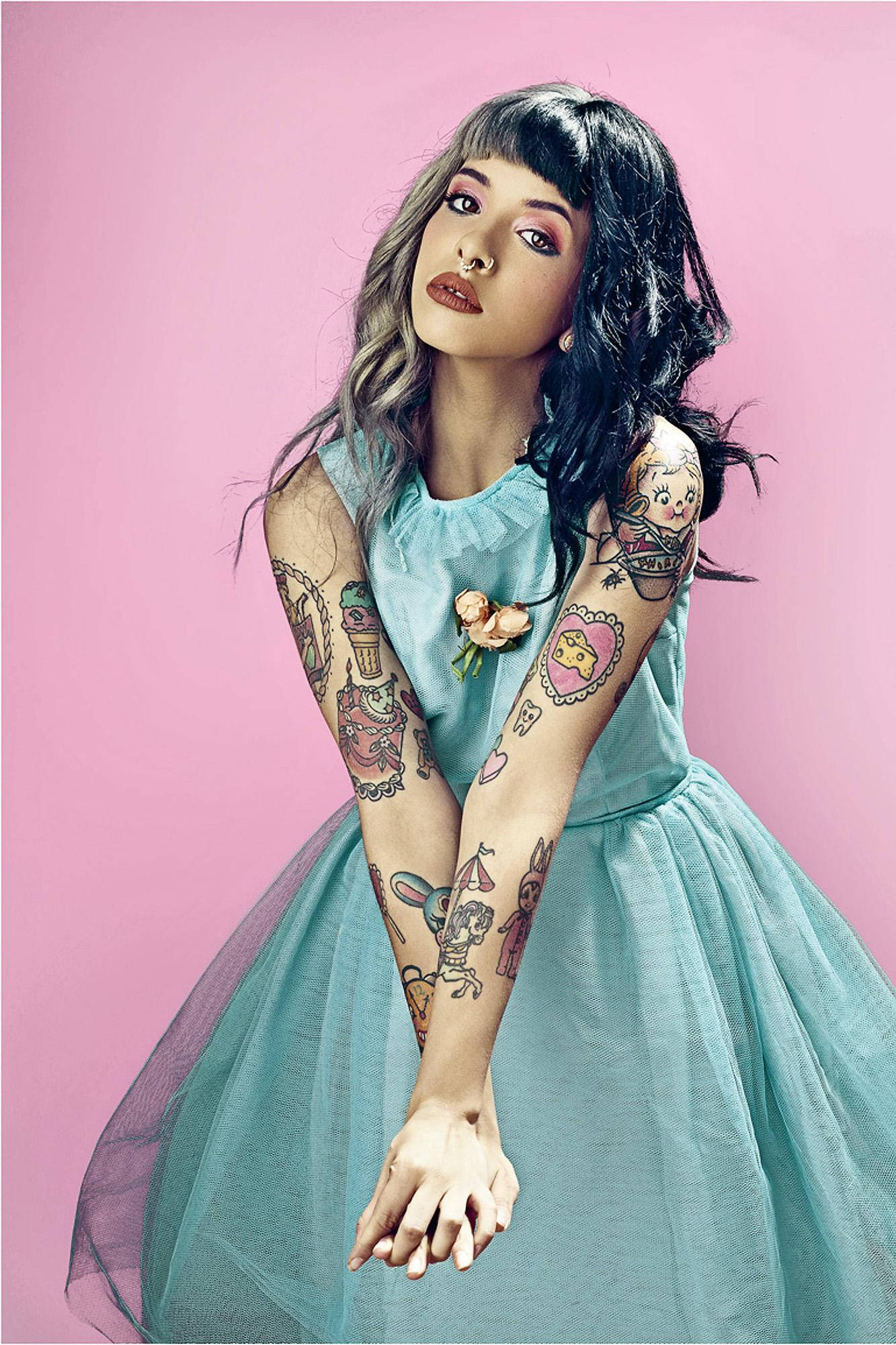 Melanie 桌布 壁紙 — 一位深色波浪髮和彩色刺青的女性，穿著淺藍色薄紗連衣裙，背景為純粉色。