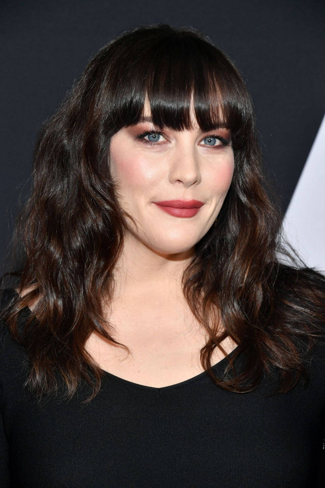 Liv Tyler 桌布 壁紙 — 一位深色波浪髮和瀏海的女性穿著黑色上衣，在深色背景前的特寫肖像。
