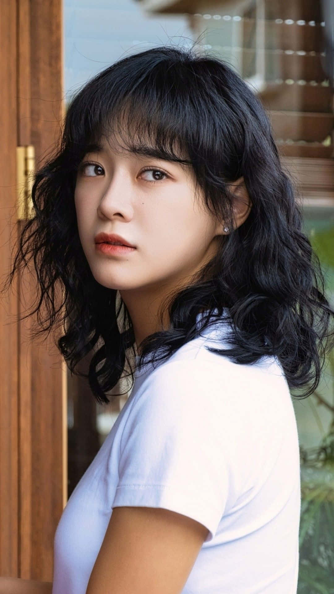 korean 桌布 壁紙 — 一位深色波浪髮和瀏海的年輕女性，穿著白色T恤，在木門附近回頭看。