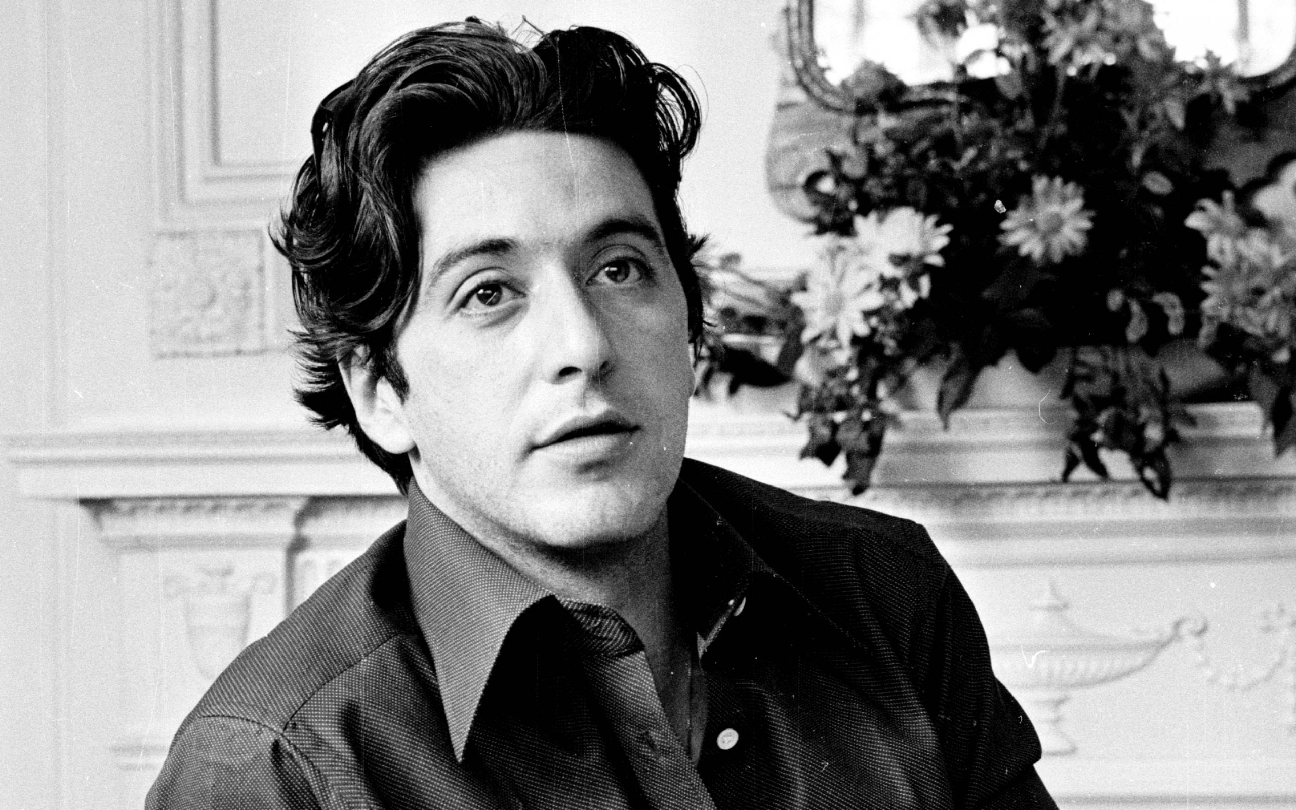 Al Pacino 桌布 壁紙 — 一位深色波浪髮年輕男子在裝飾華麗的壁爐和花卉佈置的正式房間中向上看的黑白肖像。