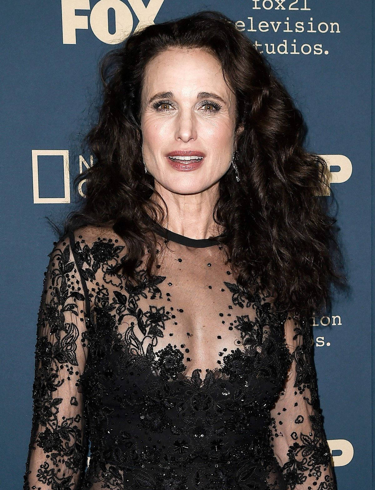 andie macdowell 桌布 壁紙 — 一位深色波浪髮的女性在FOX活動中穿著黑色蕾絲連衣裙，背景為藍色品牌背景。