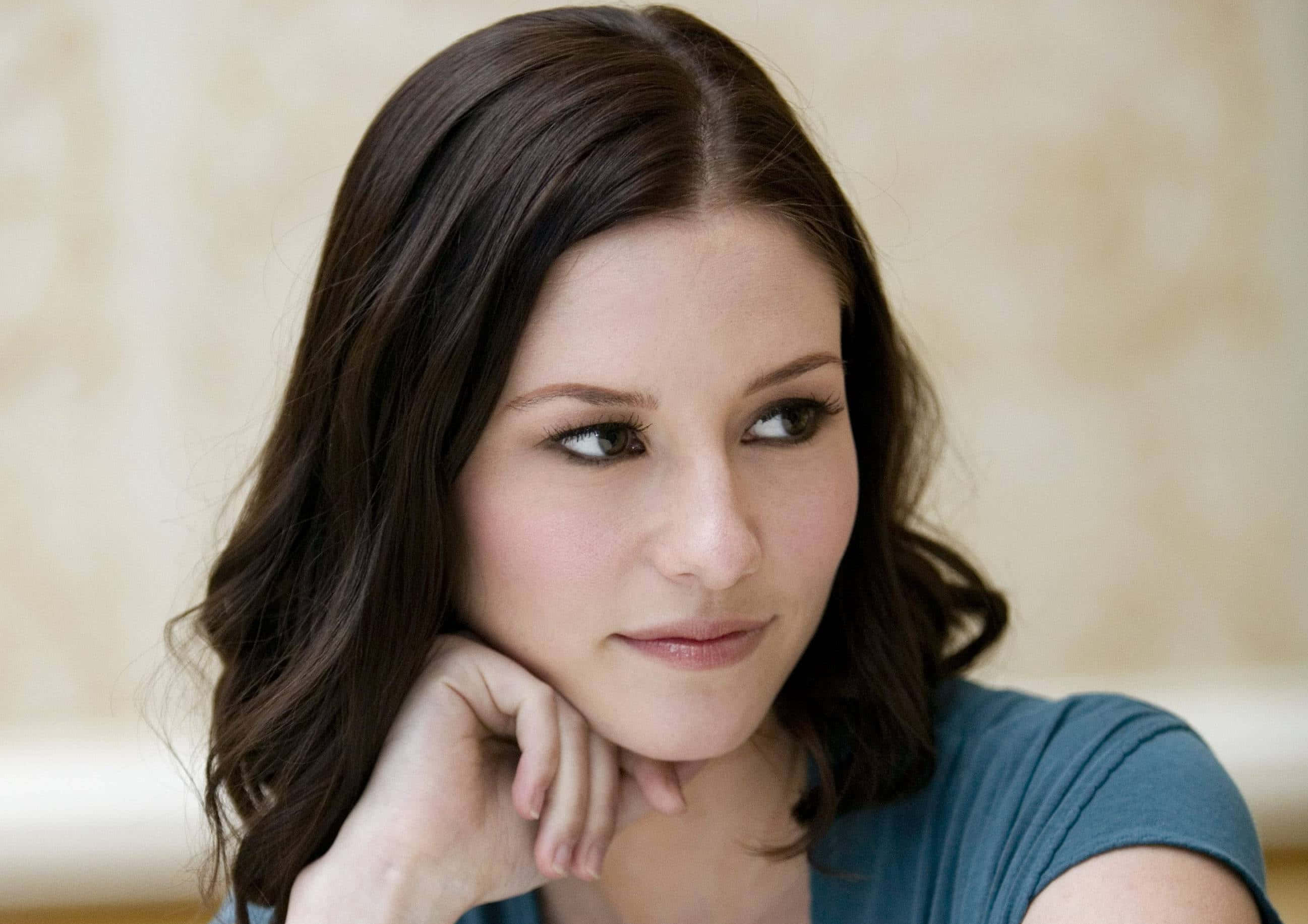 Chyler Leigh 桌布 壁紙 — 一位深色波浪髮的女性將下巴靠在手上，倚著柔軟的米色紋理牆壁。