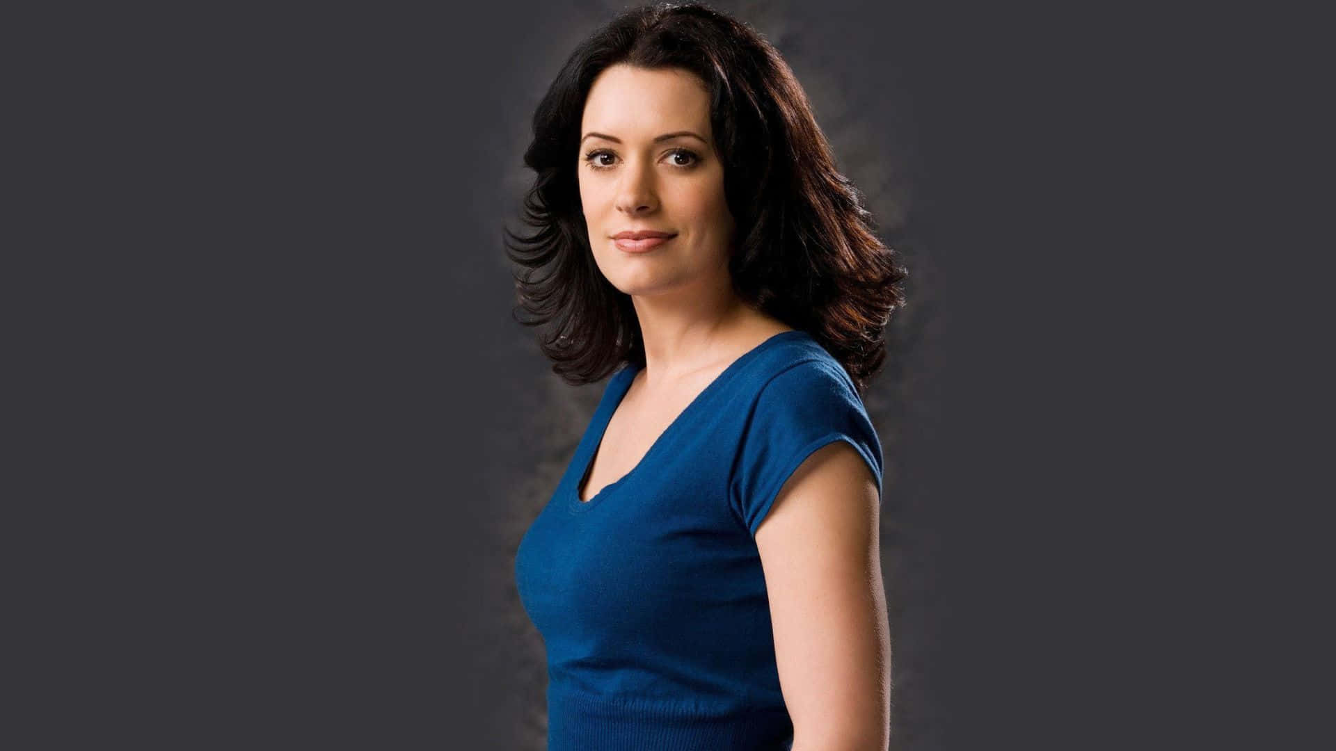 paget brewster 桌布 壁紙 — 一位深色波浪髮的女性穿著藍色V領上衣，背景為深灰色攝影棚。