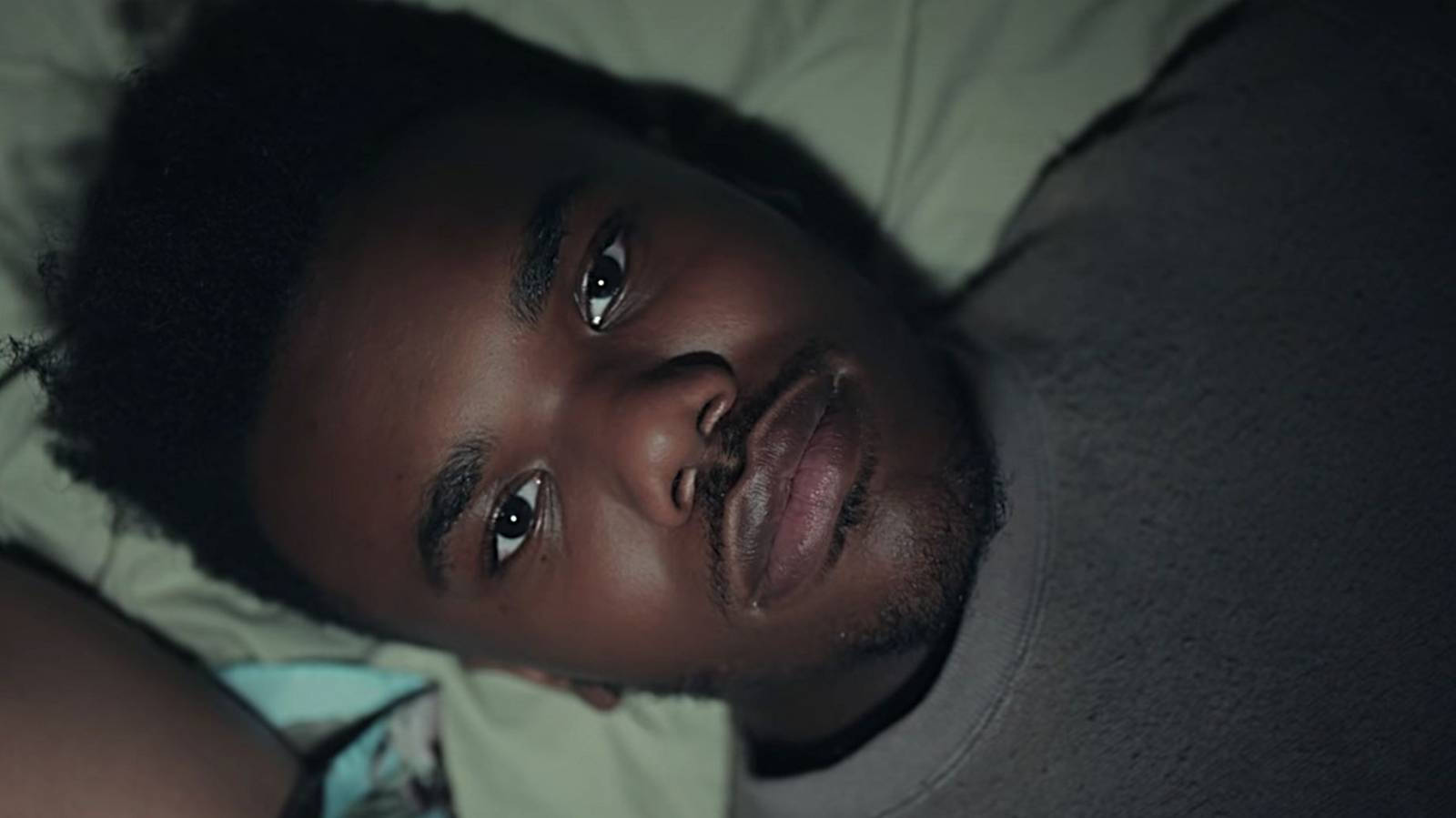 Baby Keem 桌布 壁紙 — 一位深色皮膚、捲髮的年輕男子躺在昏暗燈光下的床上，帶著沉思的表情向上凝視。